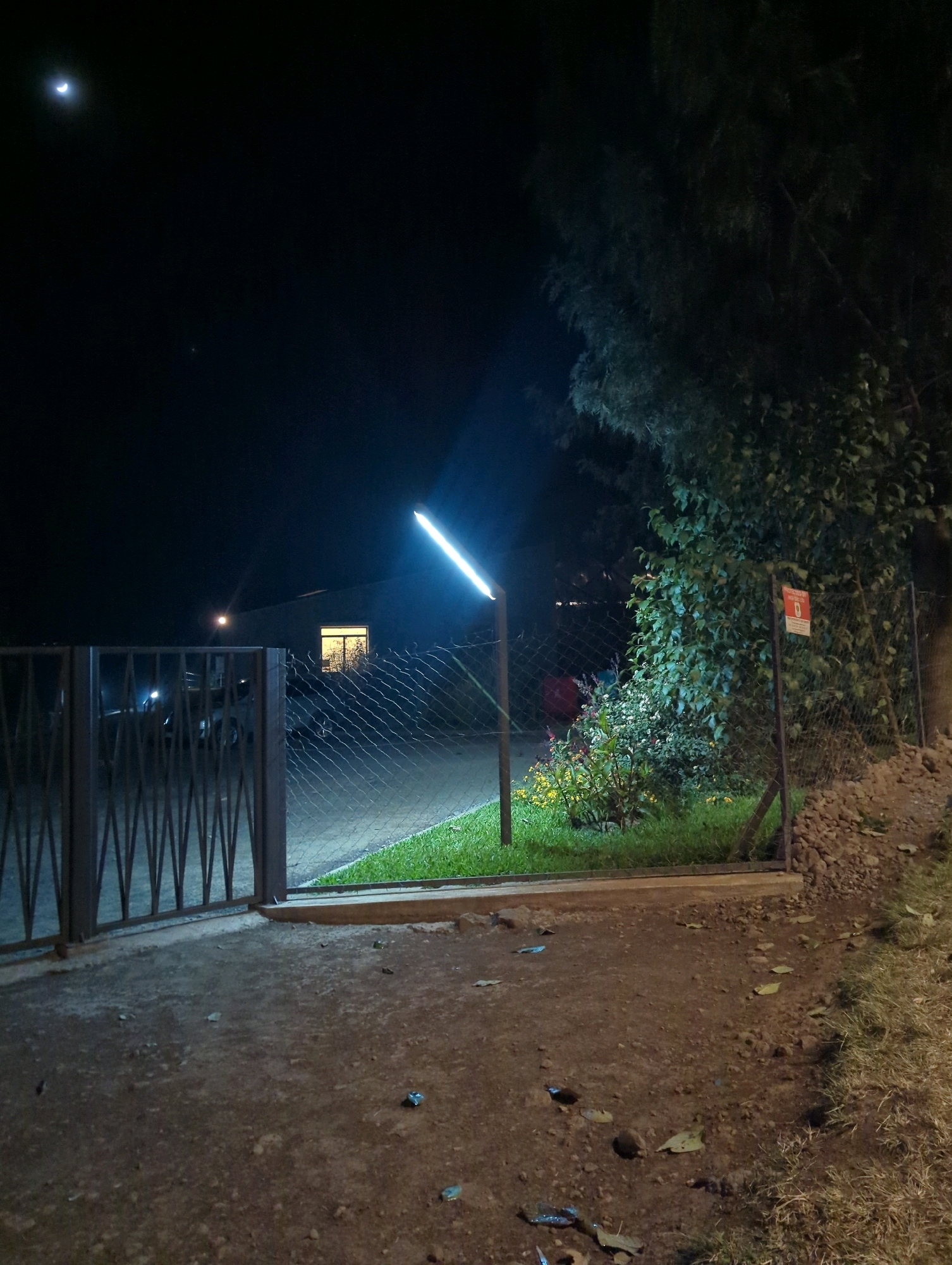 The image depicts a nighttime scene on a dirt and gravel pathway leading to a metal gate. Beyond the gate, a chain-link fence encloses a grassy area with a small garden of flowering plants. To the left, a building with a brightly lit window is visible. A modern, linear light fixture illuminates the scene. The sky above is dark, with a crescent moon visible in the upper left quadrant, along with a small, bright point of light which could be a star or planet. A large, leafy tree dominates the right side of the frame, casting shadows. A red and white sign is attached to the fence, partially obscured by leaves. The overall environment suggests a residential or semi-rural setting. The location is identified as Musanze District, Rwanda.