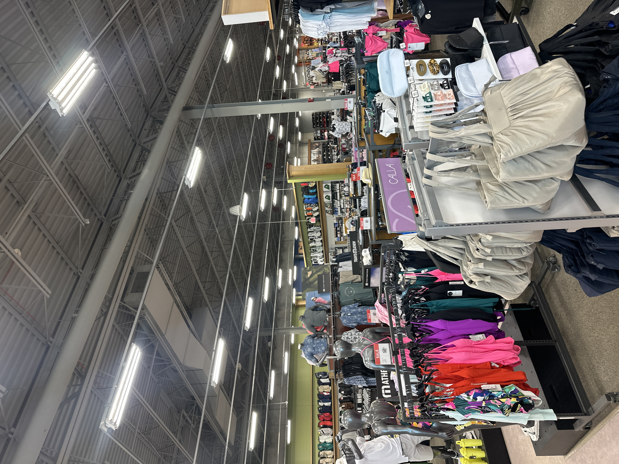 This image shows the interior of a retail store, likely a sporting goods or apparel store. The scene is well-lit, with rows of clothing racks and displays filled with various items such as swimwear, shirts, shorts, and bags. Mannequins are present, showcasing some of the apparel. In the foreground, there are racks of brightly colored swimwear and several large, neutral-colored tote bags. In the background, more clothing is displayed on racks and shelves, along with what appear to be golf bags. The store has a high ceiling with exposed industrial-style ductwork and lighting fixtures. The overall impression is that of a well-stocked retail environment. This store is located at 732 S Randall Rd, Algonquin, IL 60102, United States.