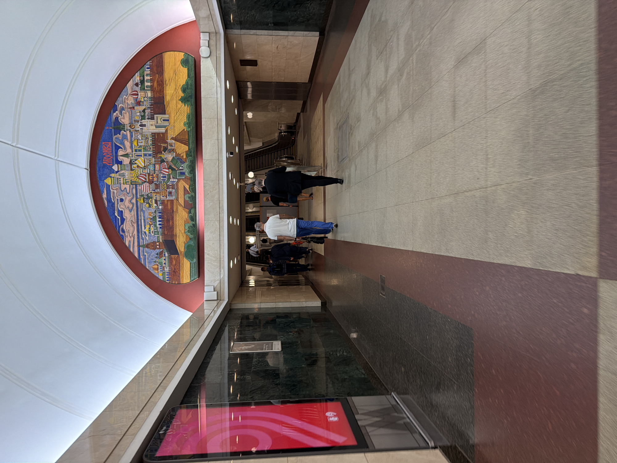 The image depicts the interior of a metro station in Moscow, Russia, likely the Novokuznetskaya station given the mosaic artwork. A large, colorful mosaic depicting a cityscape, possibly the Moscow Kremlin, adorns the arched ceiling. Several individuals are walking down a wide, well-lit corridor. The floor is a mosaic of light grey and dark red tiles, with a dark green marble wall on the left and a lighter marble wall on the right. A digital display screen is visible on the left wall, showing a bright pink graphic. In the distance, an escalator is visible, leading upwards. The overall impression is of a clean and modern public transit space, enhanced by the prominent artwork.