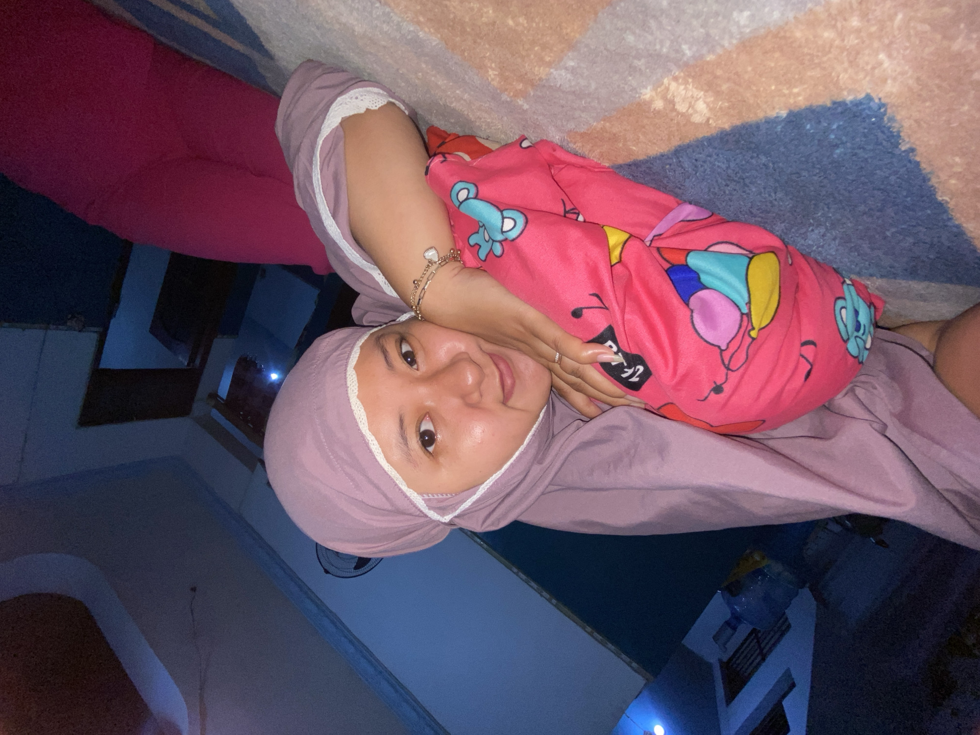 The image features a young woman, likely in her late teens or early twenties, reclining indoors, possibly on a bed or floor. She looks directly at the camera with a calm, slightly smiling expression. She has fair skin, dark eyes, and dark eyebrows.
She is wearing a light purple hijab with white lace trim around the face opening and what appears to be a matching long-sleeved garment with similar lace trim at the wrists. Her lower body is covered by bright fuchsia pink pants. A vibrant pink blanket, patterned with colorful cartoon characters like bears and balloons, covers her torso and right arm. She wears a delicate gold bracelet with a heart-shaped charm on her left wrist and a silver ring on her right ring finger.
The background includes a patterned rug with geometric designs in shades of blue, brown, and beige. The indoor setting is dimly lit, suggesting it is evening or night. A white ceiling fan is partially visible in the upper left. Through what appears to be an open window or doorway in the background, a multi-story building with visible railings can be faintly seen under the dark sky, along with a large water dispenser bottle. The overall mood is relaxed and comfortable. No discernible text is visible in the image. The location is Gelam, Indonesia.