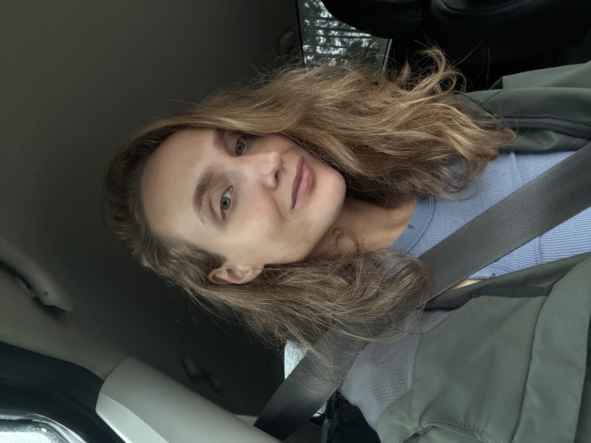 A young Caucasian woman with long, wavy, light brown hair and blue eyes is pictured from the chest up, sitting in the driver's seat of a car. She is wearing a light blue ribbed shirt and a dark green jacket. A gray seatbelt is fastened across her body. The interior of the car is visible, including part of the dashboard and the window. Outside the window, trees are visible, suggesting a natural or semi-rural environment. The lighting suggests it is daytime. The woman is looking directly at the camera with a slight smile. The background is dark and out of focus.