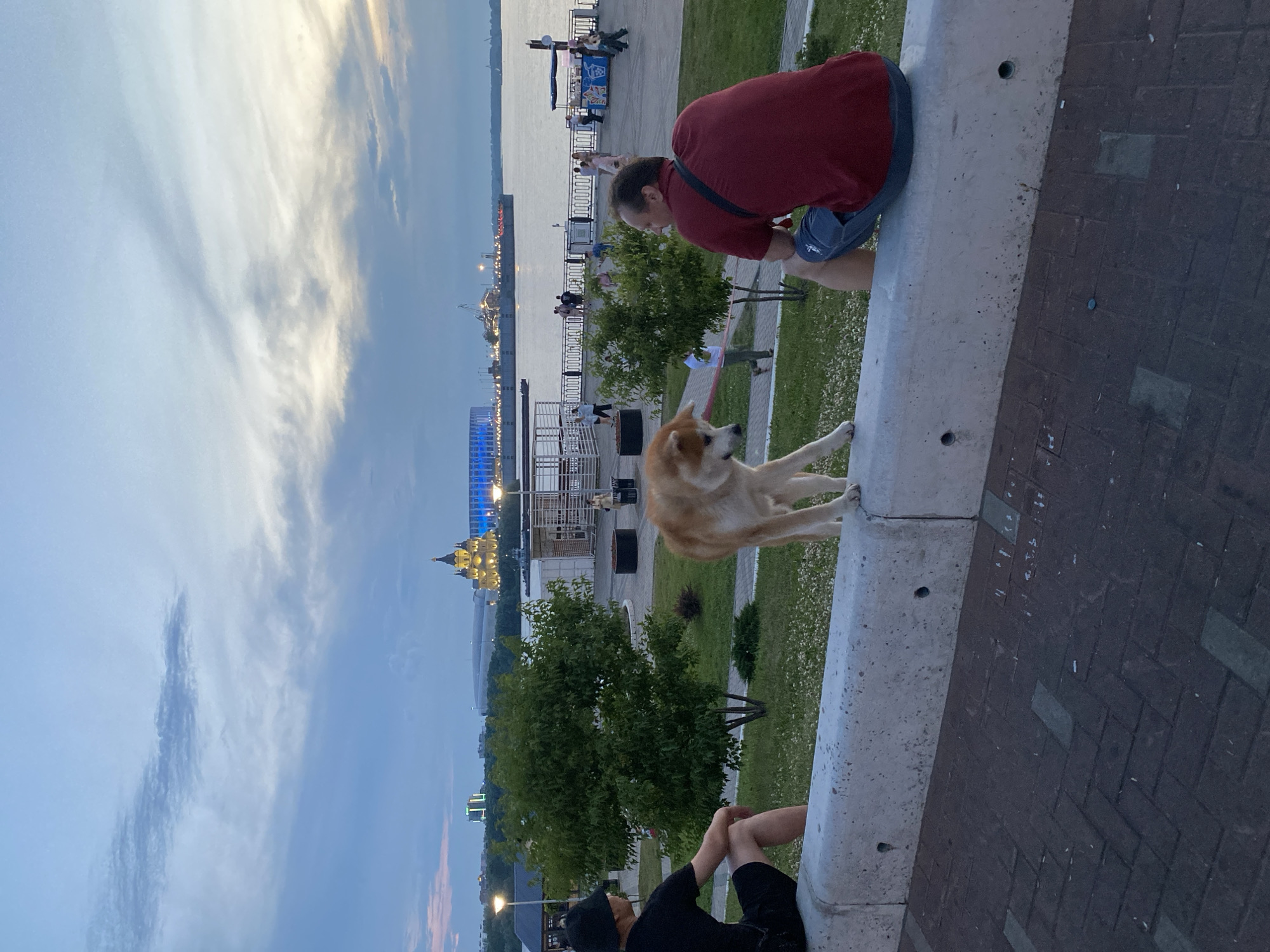 In Nizhny Novgorod, Russia, on a riverfront promenade, two men and a dog are the primary subjects. The man on the left, wearing a black hat and black t-shirt, sits on a concrete barrier facing away from the camera. To his right, a light brown dog stands on the same barrier, looking down. On the right side of the frame, a man in a red t-shirt and blue shorts also sits on the barrier, facing away from the camera and to the right.
In the background, a wide river is visible, with boats and a cityscape on the opposite bank. A prominent golden-domed building, likely the Annunciation Cathedral, is illuminated, and a large modern stadium with blue lights is also visible. Several other people are scattered along the promenade, some walking and others gathered near food stalls. The sky is partly cloudy, with a mix of blue and grey hues, suggesting it is either late afternoon or early evening. The ground in the foreground is paved with dark bricks. The overall atmosphere is relaxed, with people enjoying the evening by the river.