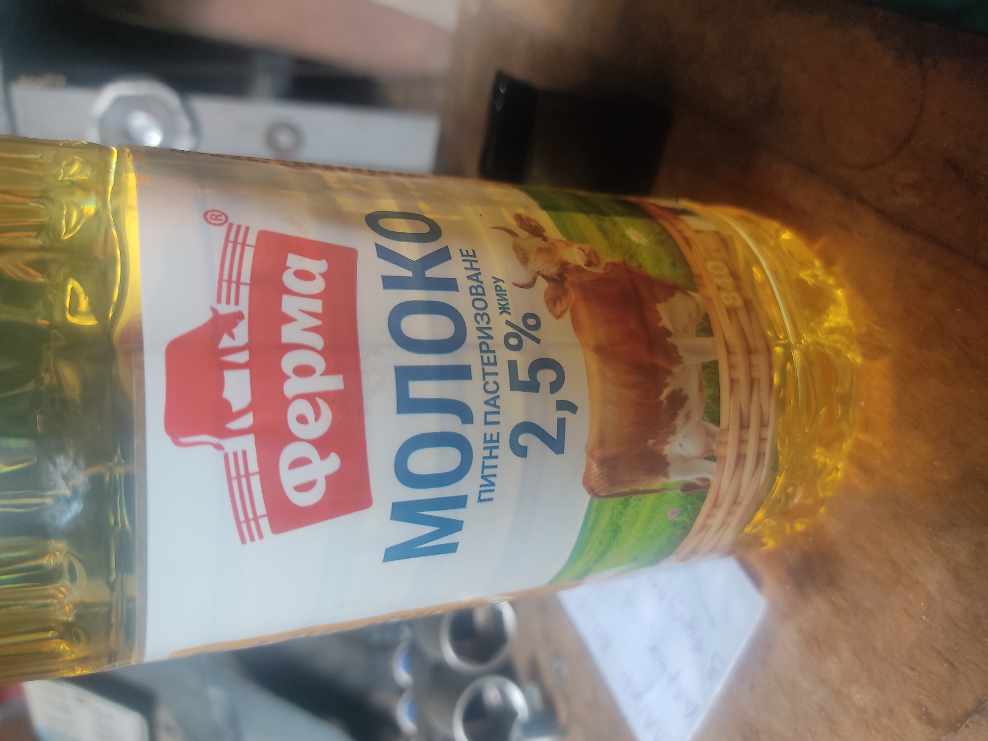 The image shows a bottle of "Farma" brand milk with 2.5% fat content. The label is predominantly white with blue and red lettering. The word "МОЛОКО" (milk in Cyrillic) is prominently displayed in large blue letters. Below it, "ПИТНЕ ПАСТЕРИЗОВАНЕ" (drinking pasteurized) is written in smaller blue letters, followed by "ЖИРУ 2,5 %" (fat 2.5%). To the left of "МОЛОКО," the brand name "Ферма" is in red. A red outline of a cow's head is part of the logo. A pastoral scene with a cow grazing in a field is depicted on the right side of the label, framed by a woven pattern. The bottle appears to be a typical plastic milk container. The liquid inside is a pale yellow, suggesting it might be oil rather than milk, which would typically be white.
The bottle is placed on a wooden surface, possibly a table. The background is out of focus, showing hints of other objects and a blurred indoor environment. The lighting suggests it might be daytime, with artificial light also present. The image is taken from a slightly high angle, looking down at the bottle. The orientation of the bottle is tilted, making the label appear angled.
Visible text includes:
- Ферма
- МОЛОКО
- ПИТНЕ ПАСТЕРИЗОВАНЕ
- ЖИРУ 2,5 %
- 840
The context provided indicates the location is Voludryntsi, Ukraine.