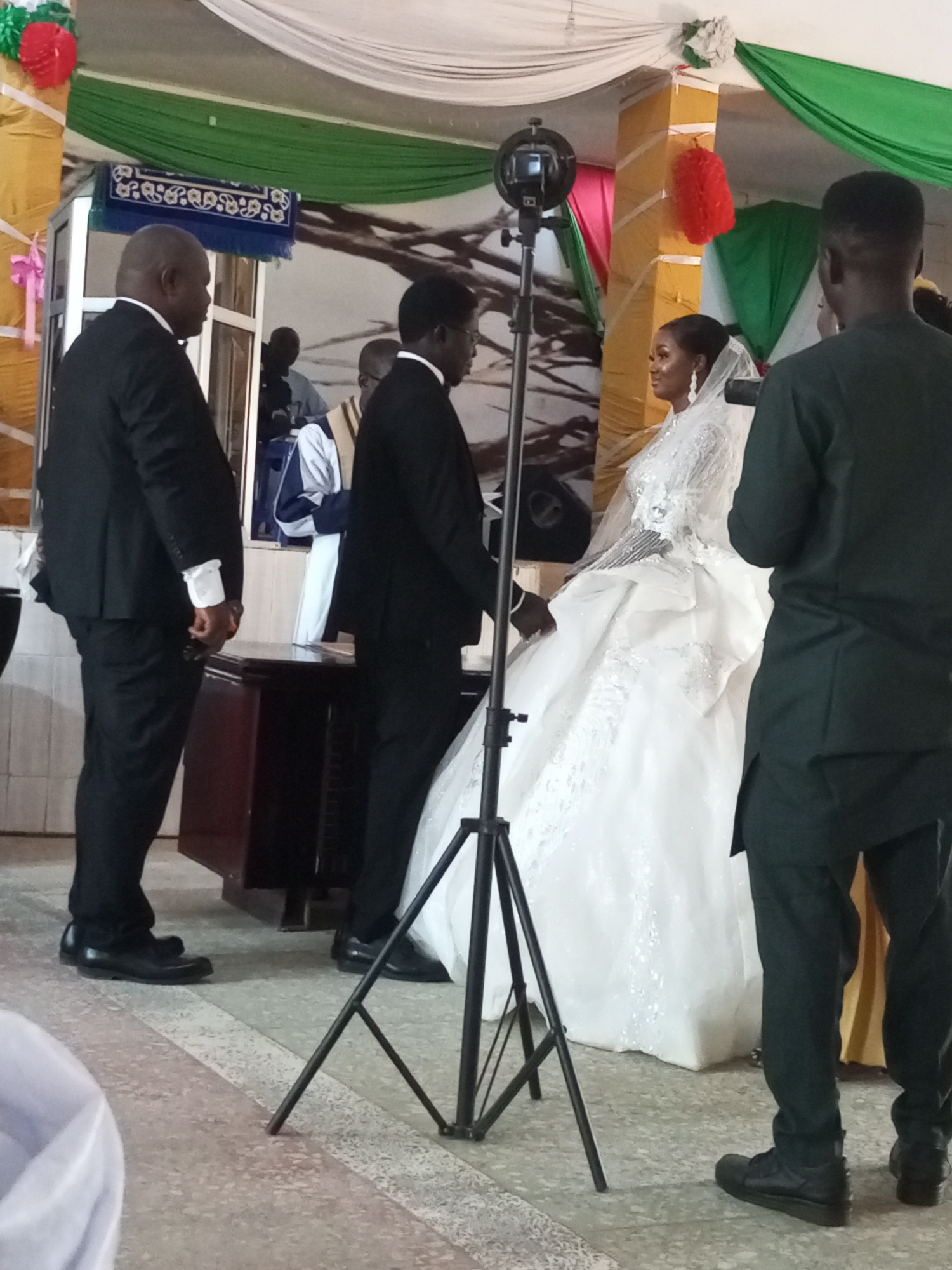 A wedding ceremony is taking place. The bride, wearing a white gown and veil, stands facing the groom, who is in a black suit. They are surrounded by two men dressed in black suits. A camera on a tripod stands in front of them. The wedding party is on a stage decorated with green, white, red and yellow decorations. The location appears to be indoors. The city and state are unknown.
