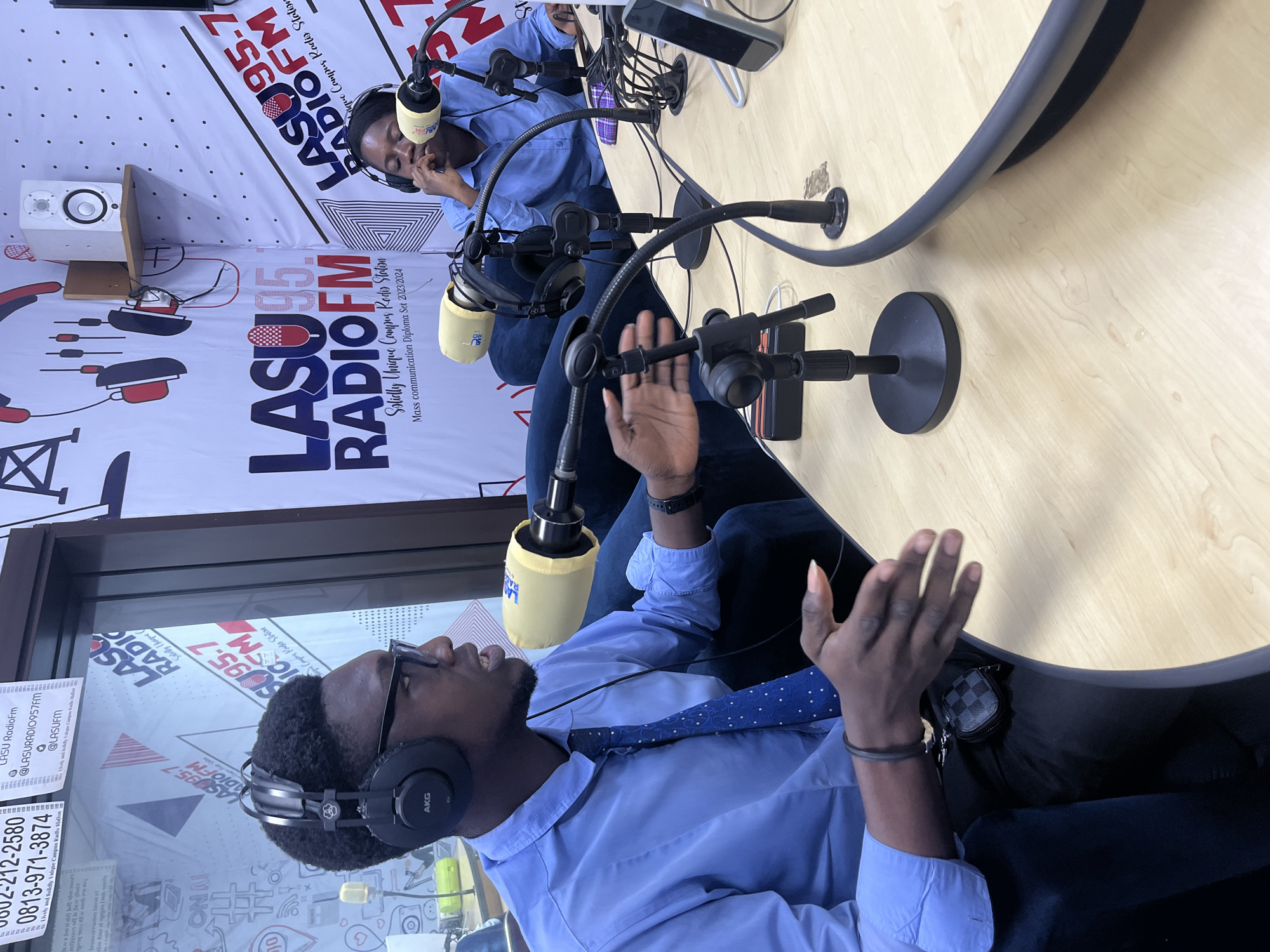 The image depicts an indoor scene within a radio studio, likely located at a university campus in Ijanikin, Nigeria, given the prominent "LASU RadioFM" branding. Two young men are seated at a curved, light wooden desk, engaged in what appears to be a broadcast or recording session.
The man in the foreground wears a light blue button-down shirt, a dark blue polka-dotted tie, dark-rimmed glasses, and black AKG headphones. His right hand is raised with an open palm, suggesting he is actively speaking or making a point. His expression appears animated and engaged. Behind him, partially visible, another man also wears a light blue shirt and headphones, with one hand near his face, seemingly listening intently.
The studio is equipped with multiple boom-mounted microphones, each fitted with a yellow foam windscreen, and professional headphones. Various cables and electronic devices are visible on the desk surfaces. In the background, a white two-way speaker rests on a shelf.
The white wall behind the subjects is adorned with banners featuring the station's branding. Visible text includes "LASU 95.7 RADIOFM" and phrases like "Solidly Unique Campus Radio Station" and "Mass communication Diploma Set 2023/2024." Contact information such as phone numbers "0802-212-2580" and "0813-971-3874" are displayed, along with social media handles like "@LASURADIO957FM" and "@LASUFM." The setting conveys a professional and active broadcasting environment, suggesting a learning or vocational context for mass communication students. The lighting is bright, indicating either daytime or well-illuminated indoor conditions.