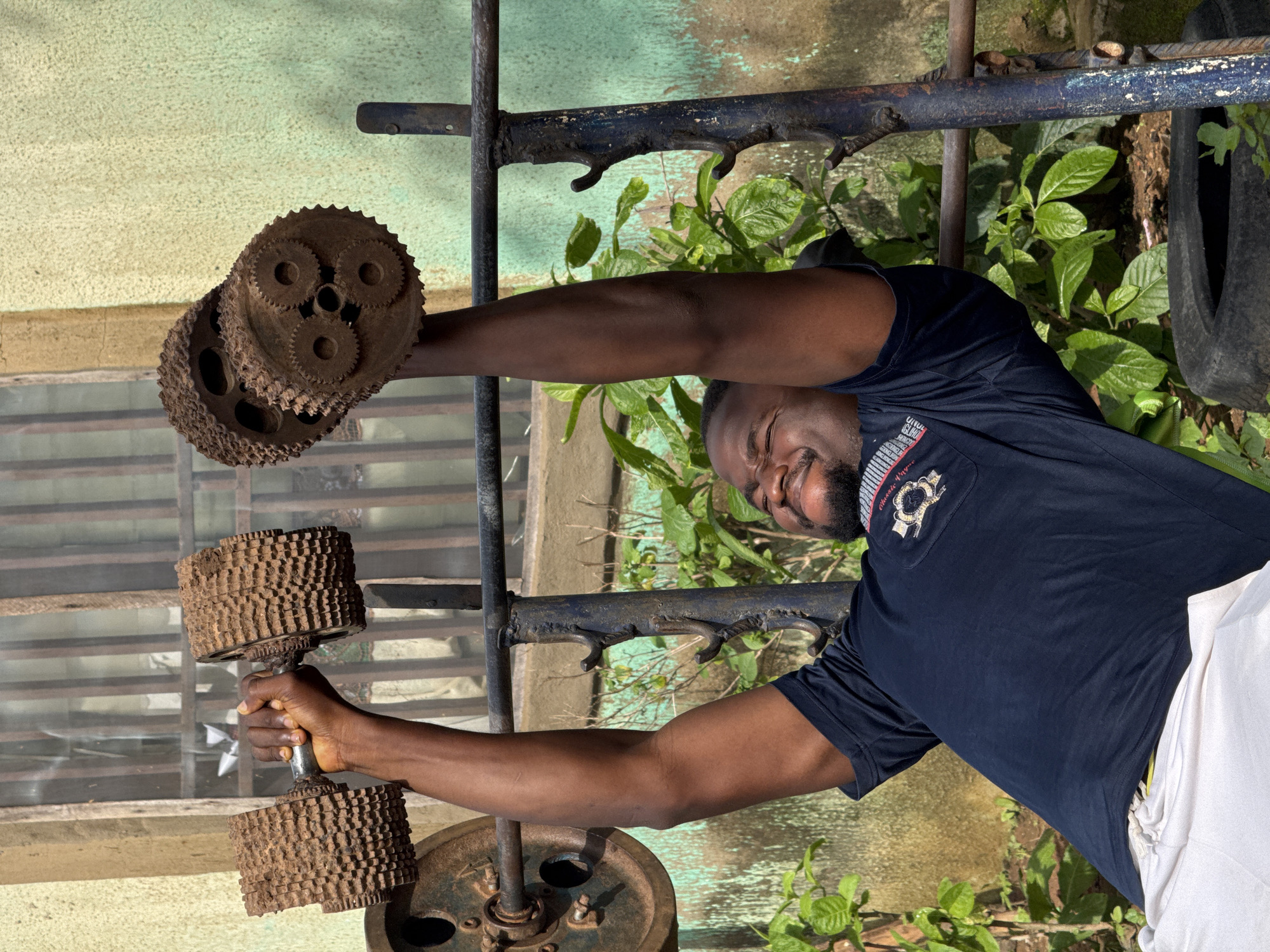 In Akwanga, Nigeria, a man is engaged in weightlifting. He is lying on a makeshift weight bench, with a barbell made of repurposed materials. The weights appear to be old gears, possibly from machinery, attached to a metal bar. The man is wearing a dark blue t-shirt and white shorts. He is in the process of lifting the barbell, with his arms extended upwards, his face showing exertion. The background suggests an outdoor setting, with a building wall, a window with bars, and lush green vegetation. The scene is rustic, implying a resourceful approach to fitness in the local environment.