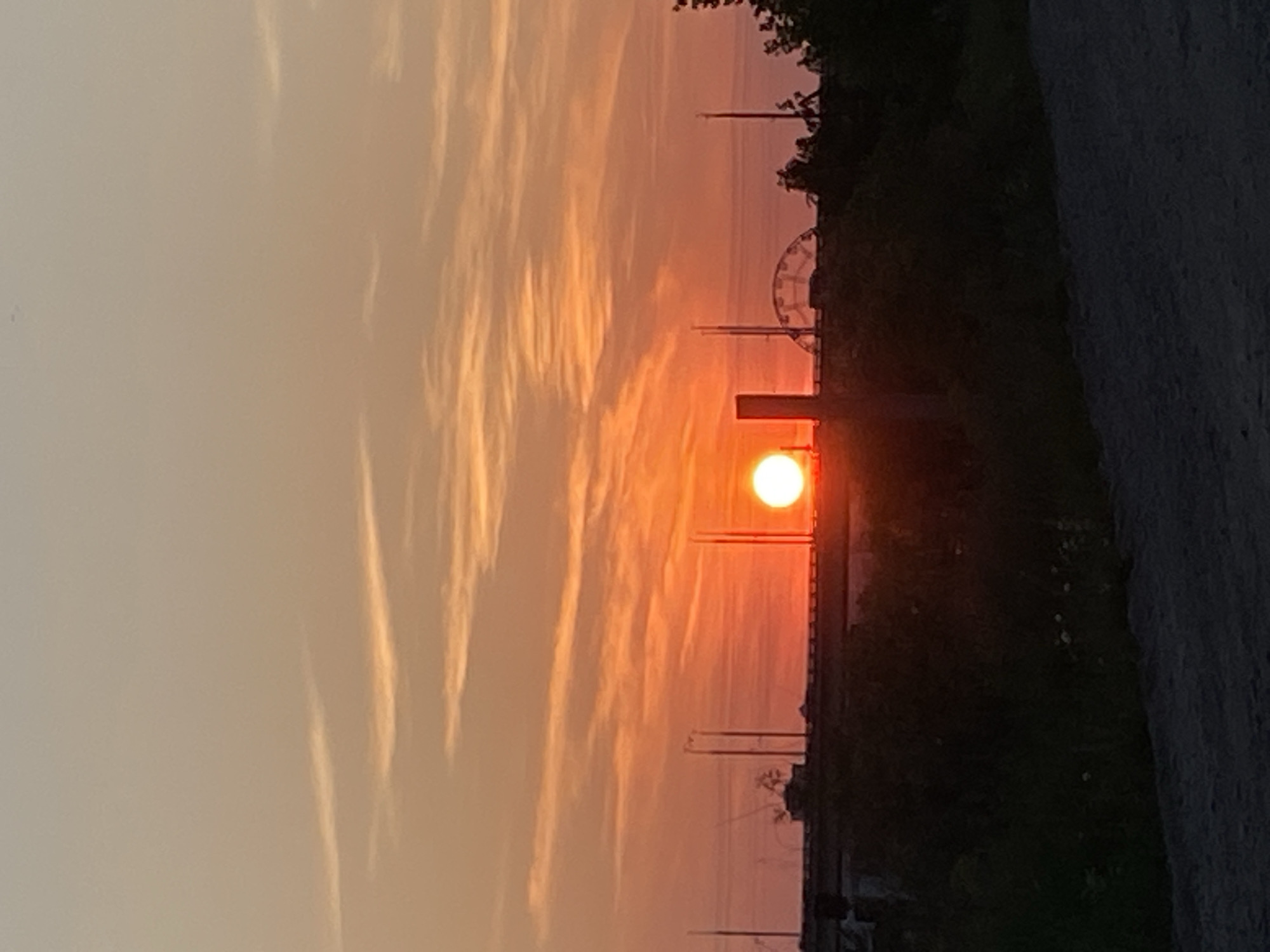 A sunset scene is depicted, with the sun being a bright orange orb near the horizon. The sky is a gradient of orange and light pink, with wispy clouds. In the middle ground, a silhouetted elevated structure spans horizontally across the frame, with a Ferris wheel-like structure visible on its left side. Several vertical poles are also present on the elevated structure. The foreground consists of dark, silhouetted vegetation and a rough, dark ground surface. The location is described as Unknown, Unknown.