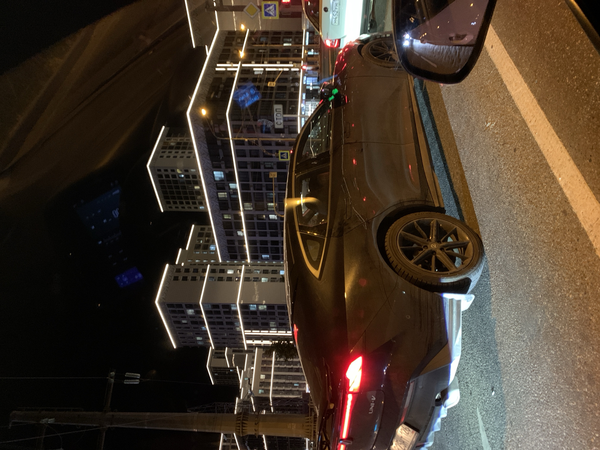 The image shows a dark-colored sedan with its taillights illuminated, seen from the rear quarter panel. The car has a sleek, modern design with a spoiler on the trunk. In the background, there are modern buildings with bright, illuminated window frames, characteristic of a city at night. A traffic light is visible, as are road markings and streetlights. The overall scene suggests a street in an urban environment at night, likely a busy road with traffic. The license plate of the car is partially visible, reading "825HT 126". The side mirror of another vehicle is in the foreground, reflecting the street and traffic lights. There are no people clearly visible in the image. The presence of the license plate format and the distinctive architecture of the buildings suggest the image might be from Russia, possibly Stavropol.