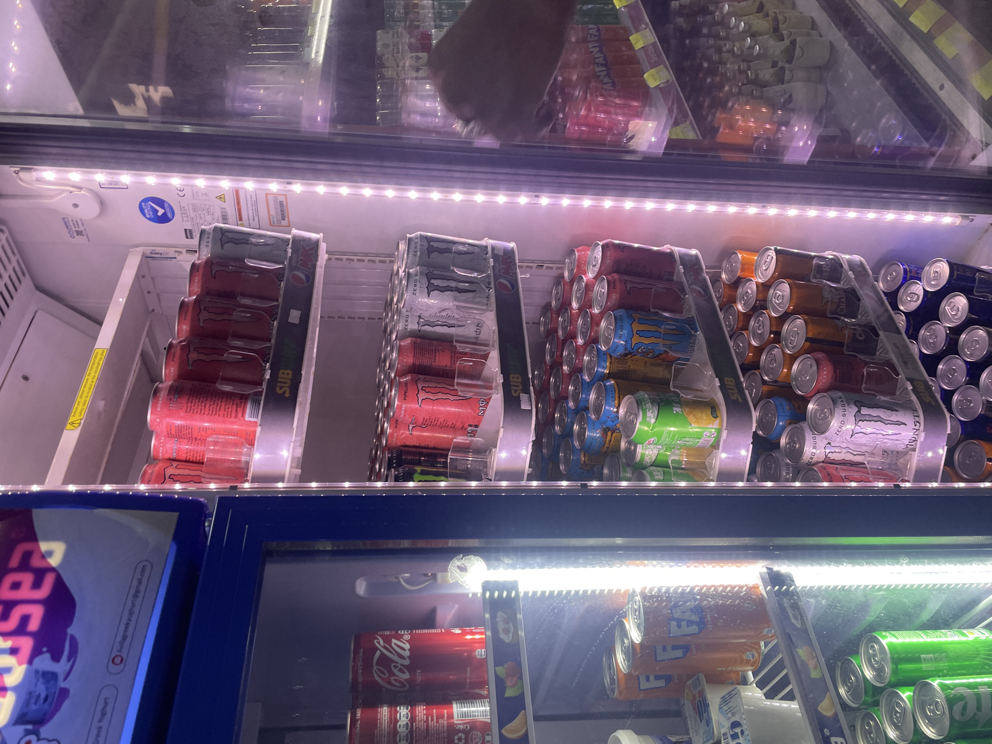 The image shows a brightly lit commercial refrigerator stocked with various canned beverages and some bottled drinks. The refrigerator is divided into multiple shelves, each displaying a selection of drinks. The left side of the refrigerator is open, revealing rows of red Coca-Cola cans and orange Fanta cans on the lower shelves. The right side of the refrigerator has glass doors and is illuminated with purple LED lights, displaying more rows of canned drinks, including red Monster Energy cans, silver Monster Energy Zero Sugar cans, and blue cans with yellow labels that read "MA YGO LOO". Bottles of drinks are visible on the top shelves of the right side. A hand is reaching into the right side of the refrigerator, suggesting someone is selecting a drink. The refrigerator appears to be in a shop or convenience store setting. The location is identified as Galadimawa Village, Nigeria, based on the provided information.