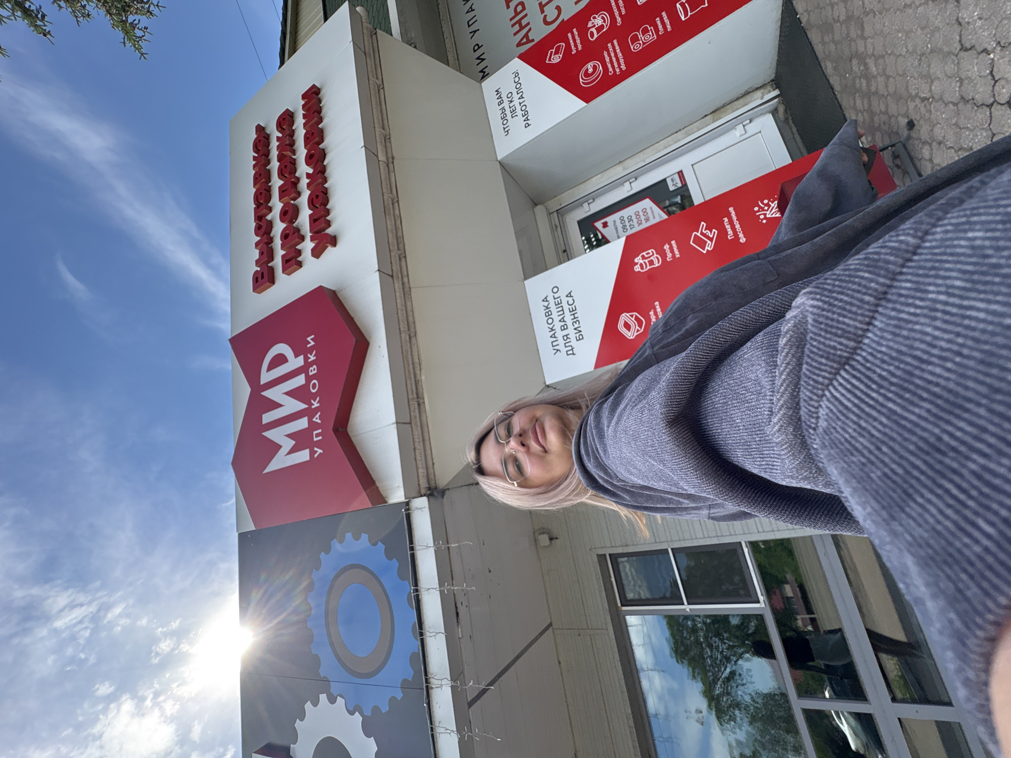 A young woman with blonde hair and glasses is taking a selfie outdoors, in front of a building with a large sign. The sign, in red and white lettering, reads "МИР УПАКОВКИ" which translates to "World of Packaging". Above this, in 3D red lettering, it says "Выставочно-продажно Упаковки", meaning "Exhibition-sale of Packaging". The building has a modern facade with large windows and what appears to be a store entrance. Red and white banners with icons and text are displayed on the front of the building, advertising various packaging products and services. The sky above is blue with scattered clouds, and the sun is visible, casting a bright glare. The ground is paved with grey concrete. The overall scene suggests a commercial establishment in an urban setting. The reflection in the window shows a street with buildings and trees.