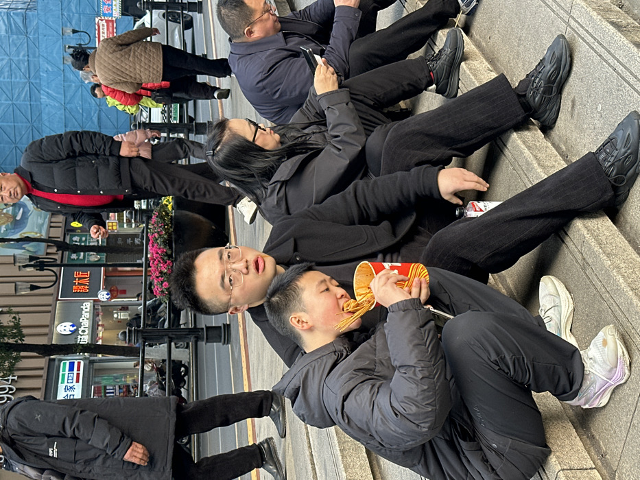 The image portrays an urban street scene in Yuzhong District, China, observed during daylight. In the foreground, four individuals are seated on concrete steps. On the left, a young male in a black puffer jacket squats on a lower step, actively eating skewered food from a red and yellow cup, with his face pressed close to the container. His cheeks exhibit a reddish tone. He leans against an adult male seated on the step above, who wears glasses, a black jacket, and pinstriped black trousers, and looks towards the right with a slightly open mouth. A small white bottle is visible near his right hand. Adjacent to him, an adult female with black glasses and a black jacket is seated, focused on a mobile phone held in both hands. To her right, a fourth adult male, also in glasses and dark clothing, is seated and engaged with a device in his lap.
Behind the seated group, a planter with pink flowers separates the steps from a pedestrian walkway and a street. On the street, an older bald male in a black puffer jacket over a red shirt walks from left to right. Other pedestrians are also visible in the background. Commercial storefronts line the street, displaying signs with Chinese characters, including "ChaPanda" (茶百道) and "木瓜匠" (Mùguā Jiàng). A vehicle is partially visible in the far background, and a building covered with blue construction netting is seen further back on the right.