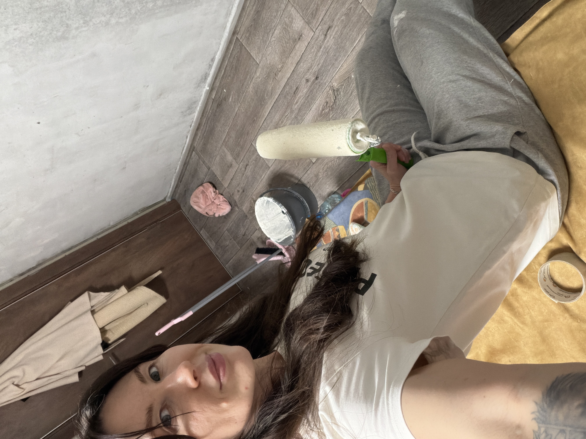 The image captures a young adult female mid-selfie, seated on a floor in a room undergoing renovation in Perm, Russia. She has long, dark hair, a slight smile, and wears a white t-shirt and grey sweatpants, with a tattoo on her left forearm. Her right hand holds a paint roller, coated in white paint or primer, with a green handle. The flooring consists of greyish-brown wood-look laminate planks. To her right, an unfinished, light-grey wall shows signs of prior use or preparation, meeting the floor with a white baseboard. On the floor nearby are a grey bucket containing white liquid, a crumpled pink cloth, and a cleaning tool with a pink-tipped handle. A roll of adhesive tape rests on a yellow surface near her left arm. A light-colored coat hangs on a dark wooden door visible in the upper left background.