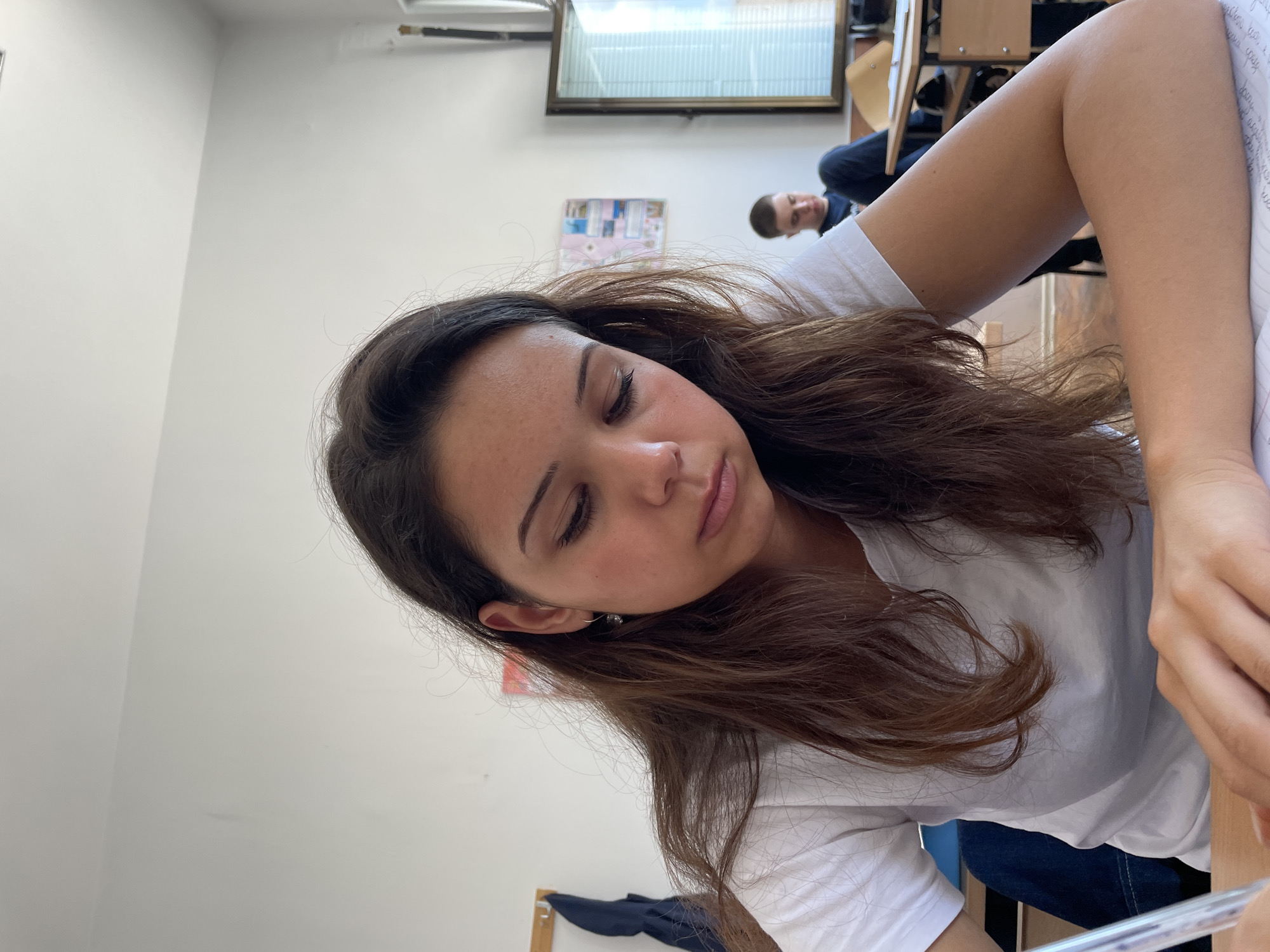 In a classroom, a young woman with long brown hair is looking down at a notebook, holding a pen in her right hand. She is wearing a white t-shirt and an earring in her left ear. In the background, another student is seated at a desk, and a window with blinds is visible. A bulletin board with papers is on the wall. The scene appears to be in a school in Zlatograd, Bulgaria.