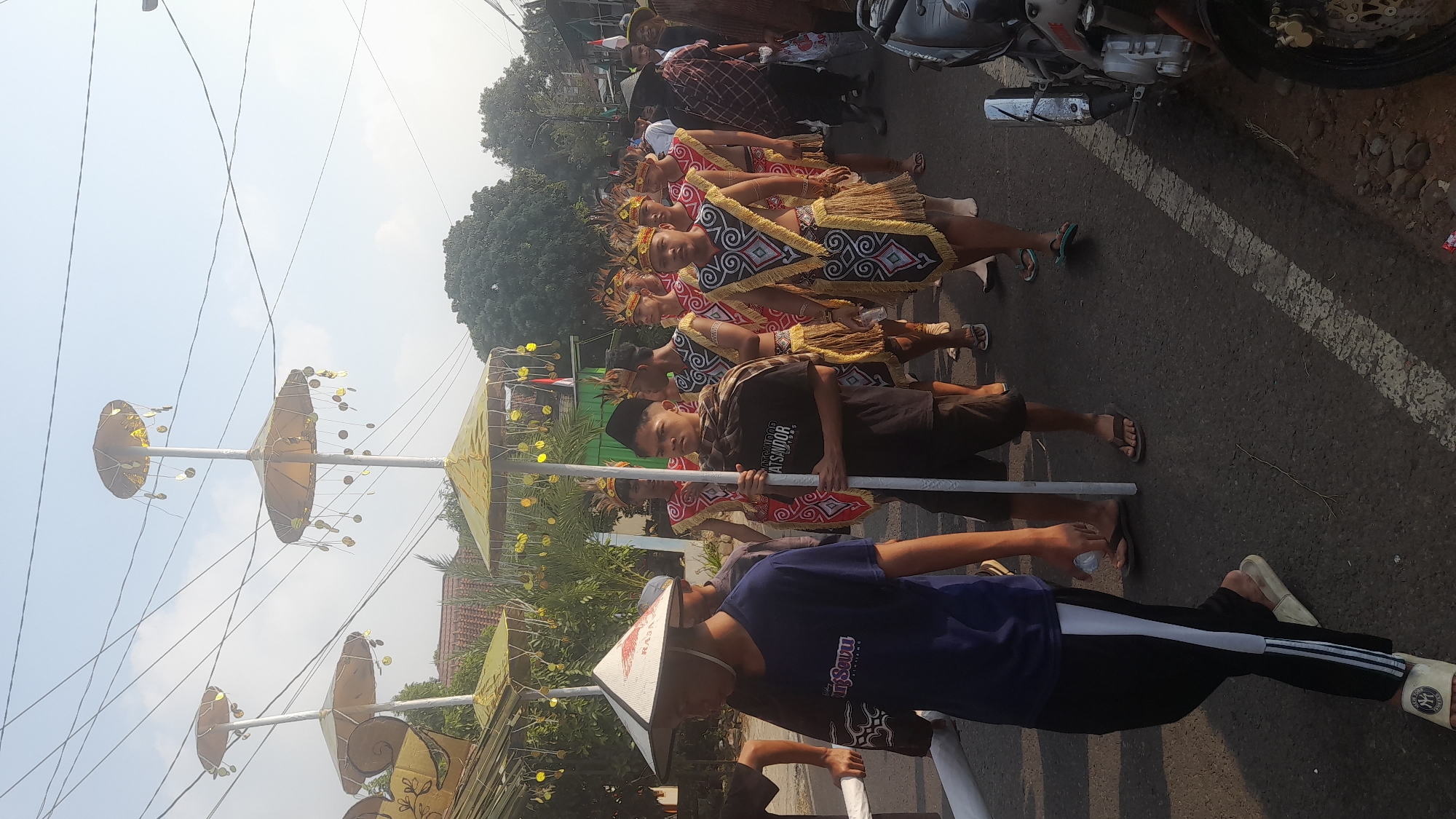 The image shows a procession in Lemahabang, Indonesia. In the foreground, a young person wearing a conical hat and a blue t-shirt walks on a street. They are followed by a group of young men in traditional attire with feathered headdresses, ornate vests, and loincloths, some carrying large, decorative parasols. The street is paved with asphalt and has white markings. To the right, a motorcycle is parked. In the background, houses, trees, and utility poles are visible under a bright, hazy sky. The event appears to be a cultural or religious celebration.