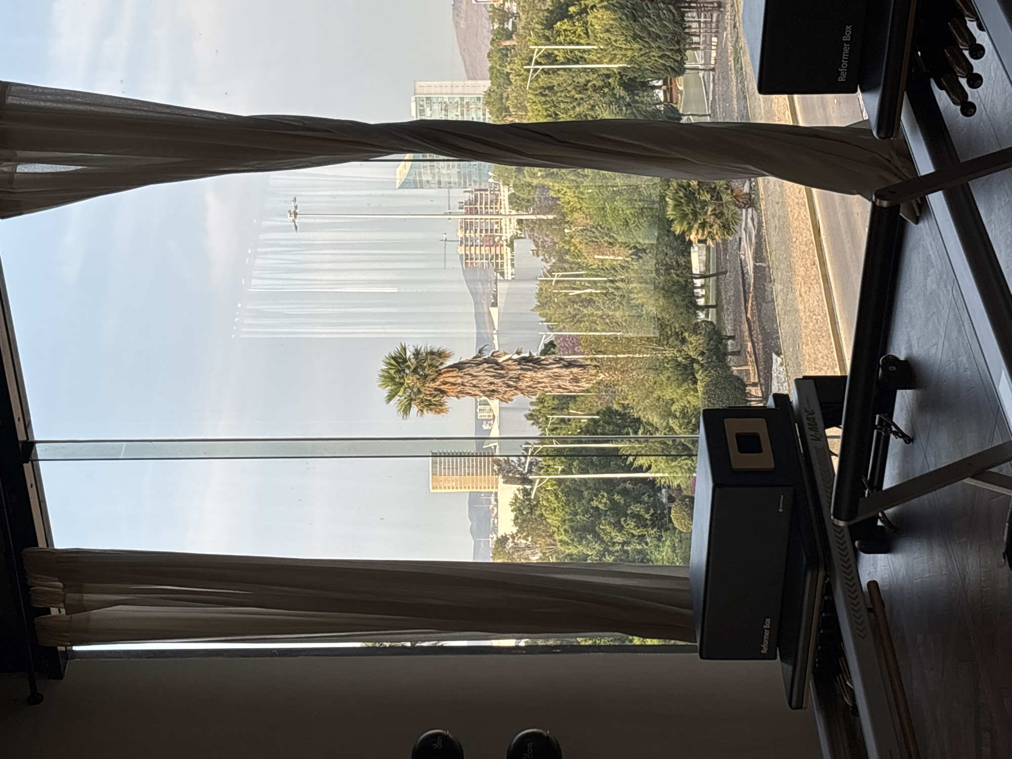 The image presents a bright daytime view from indoors, looking out through a large window. The perspective is from a room with a dark wooden floor, where the tips of a person's shoes are visible at the very bottom. On the right side of the room, a black Pilates reformer machine is positioned facing the window. A black "Reformer Box" rests on top of the reformer, and the text "V-Max" is visible on the side of the machine. Light-colored drapes frame the window on both the left and right sides, partially obscuring the view.
Outside the window, a vibrant urban landscape unfolds under a clear, bright sky. A paved road or street is visible, bordered by green foliage and numerous trees, including prominent palm trees. Several multi-story buildings, some with modern glass exteriors and others appearing more traditional, are interspersed among the greenery. In the distance, gentle hills or mountains are visible, indicating a varied topography. Reflections are noticeable on the window glass, particularly in the upper right.
The scene depicts a calm, sunny day, likely mid-morning to late afternoon, with no clouds in the sky. The combination of modern architecture, lush greenery, palm trees, and distant hills aligns with the provided location context of San Bernardino Tlaxcalancingo, Mexico. No specific activity is occurring beyond observing the outdoor scene.