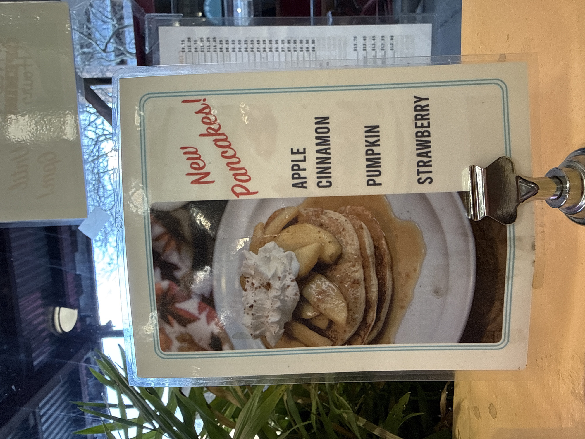 This image shows a menu advertising "New Pancakes!" The pancake flavors listed are Apple, Cinnamon, Pumpkin, and Strawberry. The image on the menu features a stack of pancakes topped with apple slices and whipped cream. The menu is held in place by a metal clip. It is unclear if the scene is in New York City.