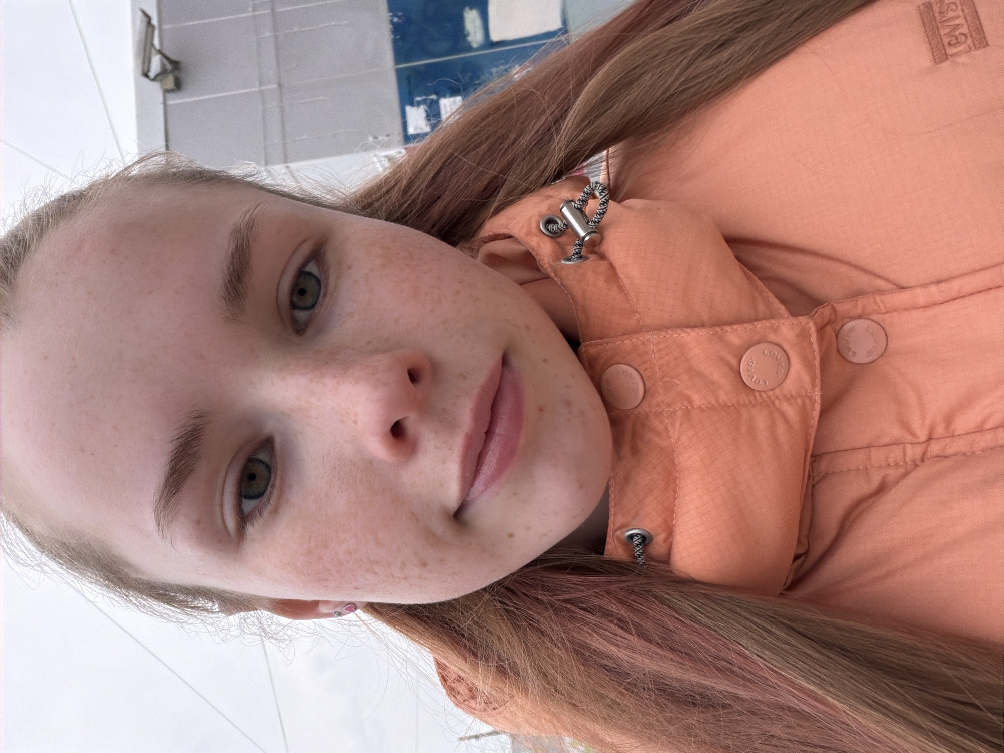 A close-up selfie of a fair-skinned young woman with light brown hair and freckles. She is wearing a peach-colored, quilted jacket with visible buttons and a drawstring clasp. The Levi's logo is discernible on the jacket. The background is blurry and appears to be a building exterior with a mix of gray and blue panels. No specific landmarks indicate the city of Berezniki, Russia.