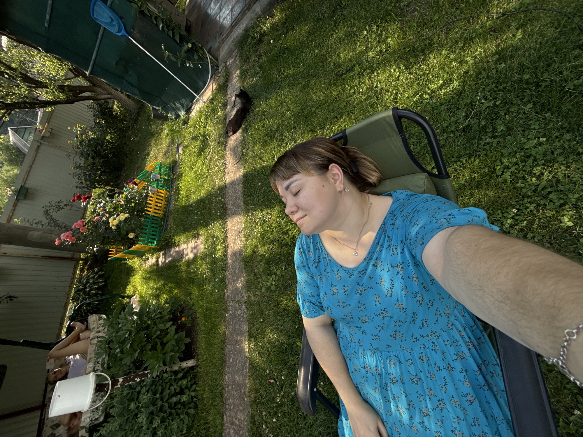 In an outdoor setting, a woman with light brown hair tied in a bun is resting in a green lounge chair. She is wearing a light blue dress with a floral pattern. Her eyes are closed, and she appears to be relaxed and sleeping, with a gentle smile on her face. A silver bracelet is visible on her wrist. The scene is bathed in the warm sunlight of what appears to be a late afternoon or early evening.
The background shows a lush green lawn with scattered white clover flowers. A stone pathway runs diagonally across the grass. To the left, there are rose bushes with pink and yellow flowers, and a colorful plastic fence. Behind the flowers, a light-colored fence and a dark green tarpaulin are visible, suggesting a backyard or garden area. In the distance, trees and foliage can be seen. A black cat is lying on the grass near the woman, also seemingly resting.
The overall impression is one of peaceful relaxation in a garden on a pleasant day. The location context provided is Nizhne-Chufichevo, Russia. There is no visible text in the image.