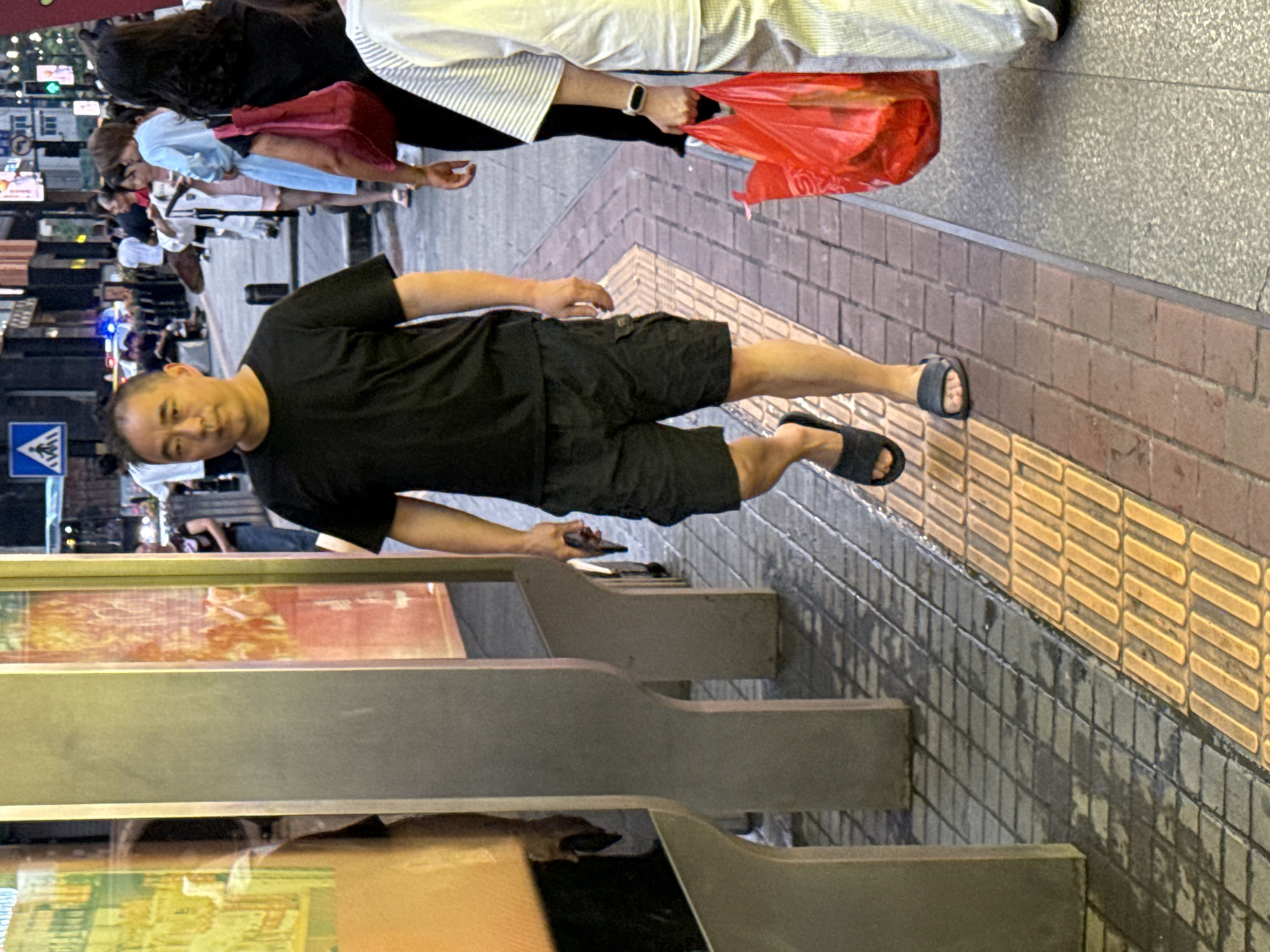 A middle-aged East Asian man with short, dark hair is walking on a city sidewalk in Yuzhong District, China. He is wearing a black t-shirt and black cargo shorts, and dark-colored slip-on sandals. The man is holding a black smartphone in his right hand. He is walking towards the viewer, slightly to the left of the frame. The sidewalk is paved with square tiles in varying shades of gray and yellow. To the left of the man are large, grey, cylindrical pillars and reflective display cases. In the background, other people are walking, and there are buildings and streetlights visible. A person in a white and striped shirt is walking to the right of the frame, carrying a large red plastic bag.