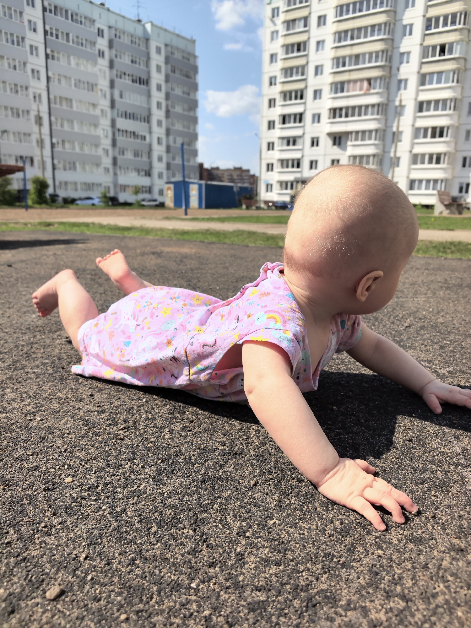 A baby is lying on their stomach on a dark asphalt surface in an outdoor setting. The baby is wearing a pink patterned dress. In the background, two tall, light-colored apartment buildings are visible. The location is in Фролы, Russia.