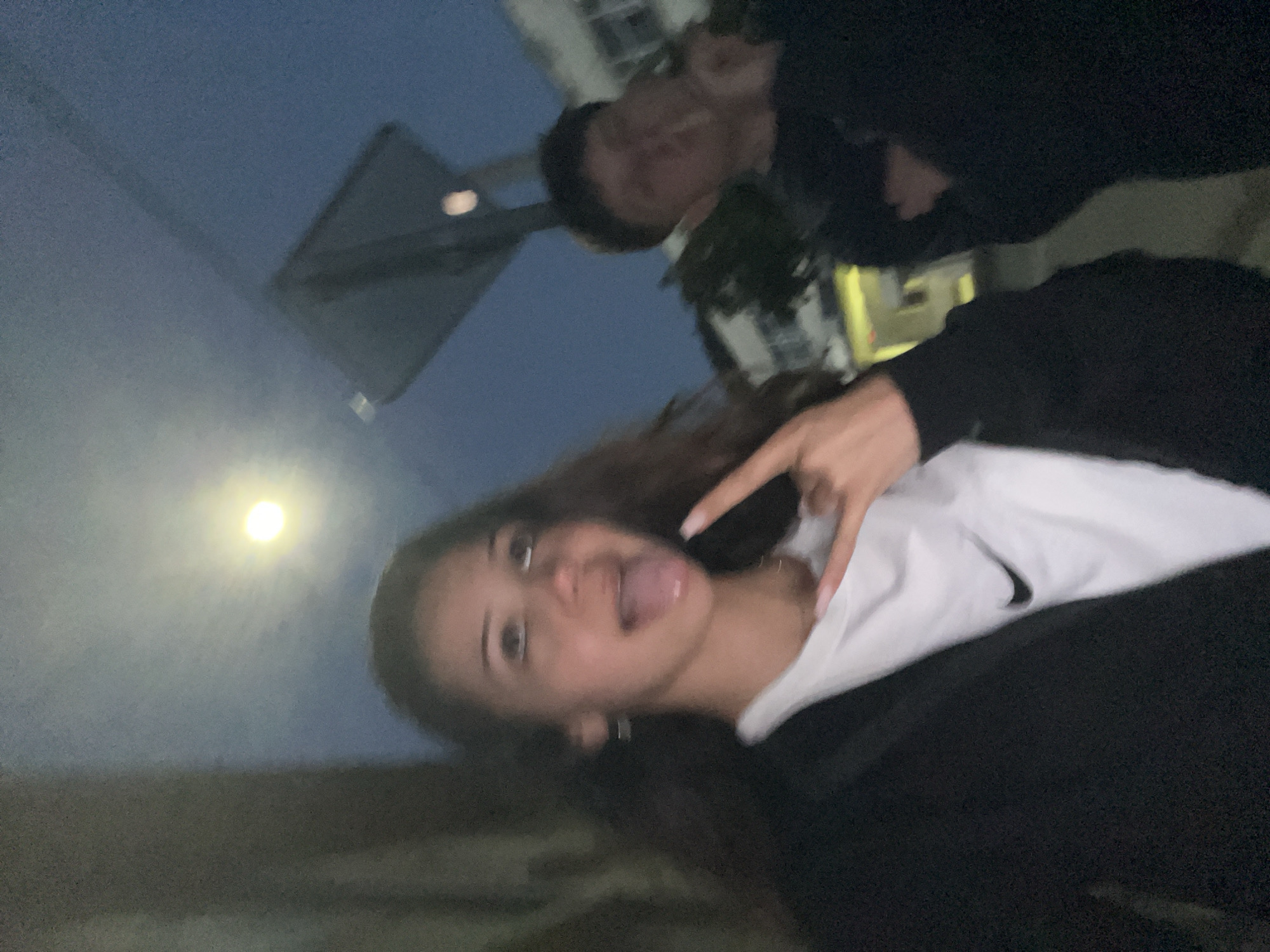 This image depicts an outdoor night scene in the city of Zlatograd, Bulgaria, featuring two young individuals. The lighting is dim, characterized by a dark blue sky and a prominent, bright spherical light source in the upper left quadrant, likely a street light or the moon. The image exhibits noticeable blur and graininess, consistent with low-light photography.
In the foreground, slightly left of center, is a young female with long, dark hair. She is wearing a black jacket or hoodie over a white t-shirt that displays a black Nike swoosh logo. Her left hand is raised to shoulder height, forming a "rock on" gesture with her index and pinky fingers extended. Her mouth is open with her tongue slightly extended, and her eyes are directed upwards and to her right, conveying an expressive, playful demeanor. She wears small hoop earrings. Her fingernails are long and light-colored.
To the right and further in the background, a young male is partially visible. He has short, dark hair and is wearing a dark-colored, long-sleeved top. His right hand is raised to his mouth, though the object or action is indistinct due to blur. He appears to be looking generally towards the left.
The background includes blurry urban elements: a dark, angular structure in the upper right, possibly a sign or part of a building, and lighter-colored buildings with windows further back on the right. Dark foliage is visible between and behind the individuals. The ground beneath them appears to be a paved surface.