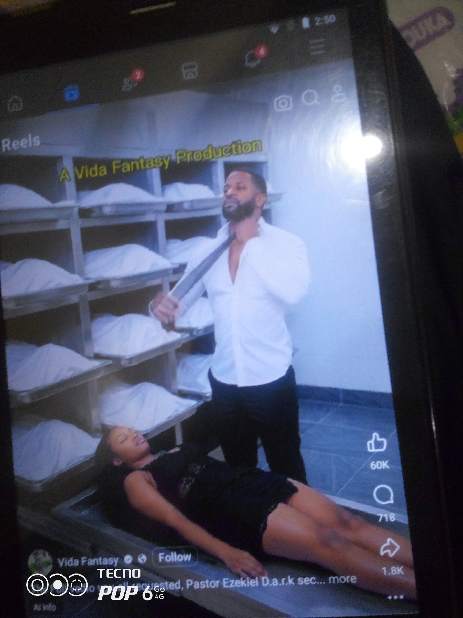 The image shows a screenshot of a video playing on a smartphone, possibly depicting a scene related to funeral services. A man in a white shirt and dark pants stands to the right, holding a tie. In the foreground, a woman is lying down, appearing to be in a morgue or similar setting. She is wearing a black outfit. The background features rows of shelves, each containing what appears to be a body bag or a similar covered item. The video's metadata indicates it's from "Vida Fantasy" and includes a "Follow" button. There are also engagement metrics like "60K" likes, "718" comments, and "1.8K" shares visible on the right side of the screen. The lower part of the screen shows "Tecno Pop 6 4G," identifying the phone model. The image does not provide any information or context about the location being Howayi, Nigeria.