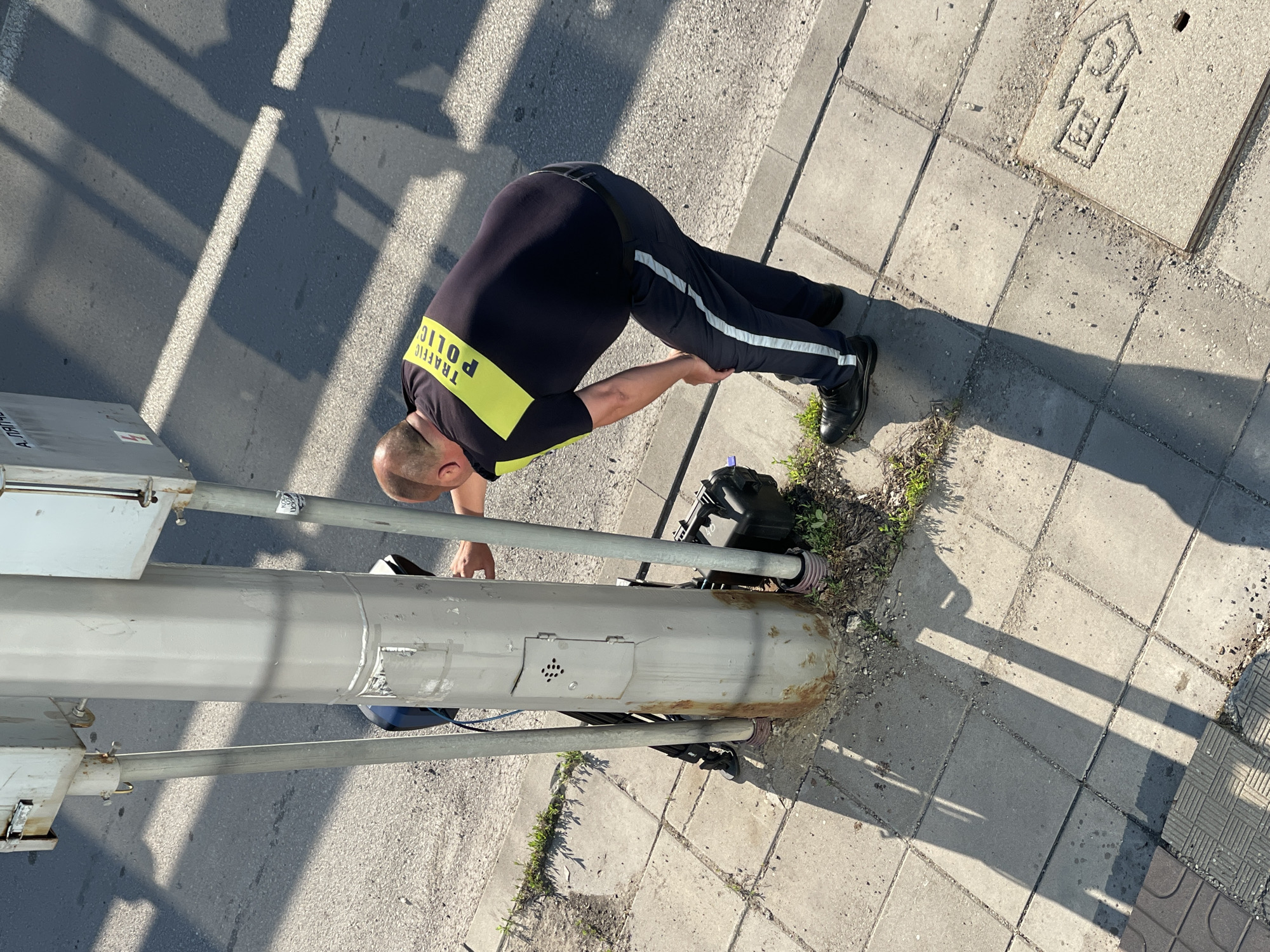 An overweight man in a dark blue uniform shirt with a bright yellow reflective stripe across the chest and a "TRAFFIC POLICE" logo on the back, and dark blue pants with a reflective stripe on the side, is bending over next to a gray metal pole. The pole appears to be a traffic signal pole. The man is wearing black shoes. He is touching or working on a black box attached to the pole with a blue wire connected to it. There is another thinner metal pole to the left of the main pole, and a black rack or structure behind them. The ground is paved with square concrete tiles, with some patches of grass and dirt. The scene appears to be outdoors, on a street or sidewalk, with shadows cast by the pole and possibly overhead structures indicating it is daytime. A gray metal box is mounted higher on the pole. The man's body language suggests he is focused on his task. The overall setting is an urban environment. The image was taken in Sofia, Bulgaria, as indicated by the city's distinctive tram track markings on the sidewalk in the lower right corner of the image, which are typically found in Sofia.