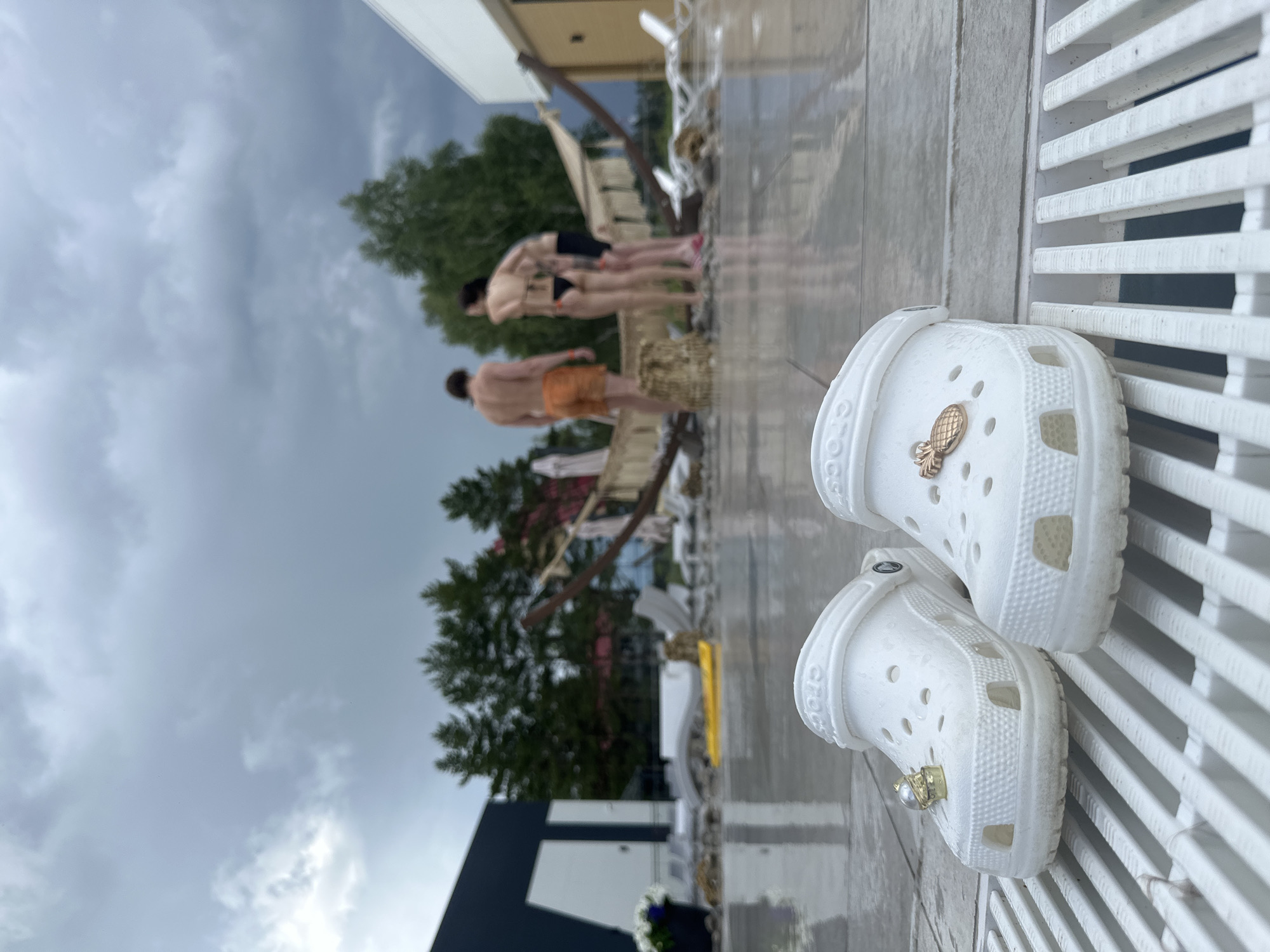 A pair of white Crocs, adorned with a gold pineapple charm on one shoe and a gold pearl charm on the other, are placed on a white slatted surface in the foreground. The Crocs are dirty, suggesting they have been worn. In the blurred background, several people are visible standing on a wet, paved area. One person is wearing orange shorts, while others are in swimwear. Trees and some outdoor furniture can be seen behind them. The sky is overcast and gray, indicating a cloudy day, possibly rainy. The overall scene appears to be an outdoor setting, perhaps a pool area or a patio, given the wet ground and swimwear. There is no clear indication of the time of day or specific location, but the weather appears somber. The Crocs are the sharpest objects in the frame, while the background is out of focus. The word "CROCS" is visible on the side of one of the shoes.