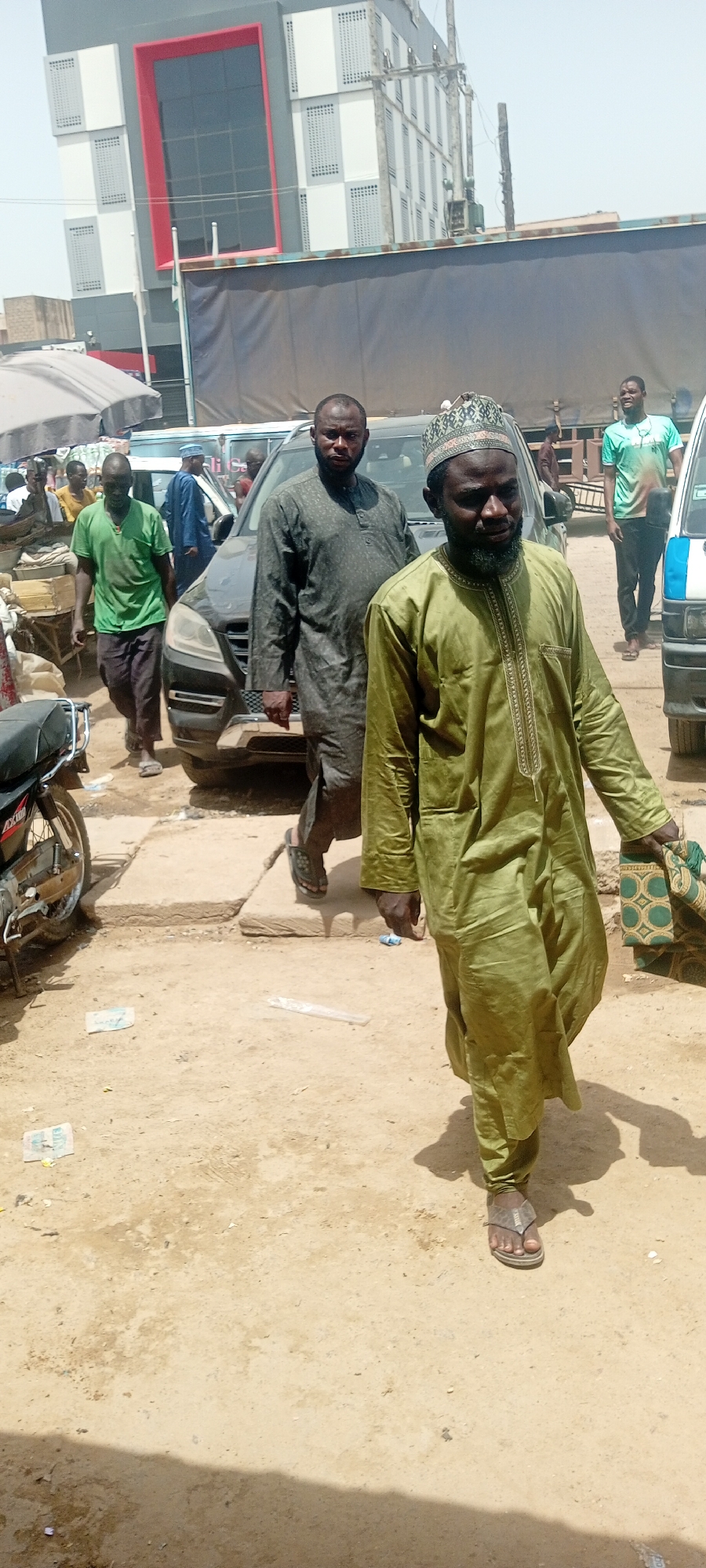 This media file depicts a street scene in Fagge C, Nigeria, likely during the daytime under clear skies. The foreground is dominated by three men walking. The man closest to the camera is wearing a long, light green traditional garment with intricate embroidery around the neck and sleeves, and a matching cap. He is carrying a patterned bag in his right hand. To his left and slightly behind him walks another man in a dark, long-sleeved garment. Further to the left, a man in a bright green t-shirt and dark pants walks away from the camera.
In the background, several vehicles are visible, including a black SUV and a white truck with a blue stripe. Behind the vehicles, a modern building with grey and white panels and a red-trimmed window section stands out. The ground is dusty and appears to be a paved or dirt road. Scattered on the ground are some pieces of paper, possibly currency. The overall atmosphere suggests a bustling market or a busy street. The men appear to be engaged in everyday activities, with no overt signs of distress or celebration.
