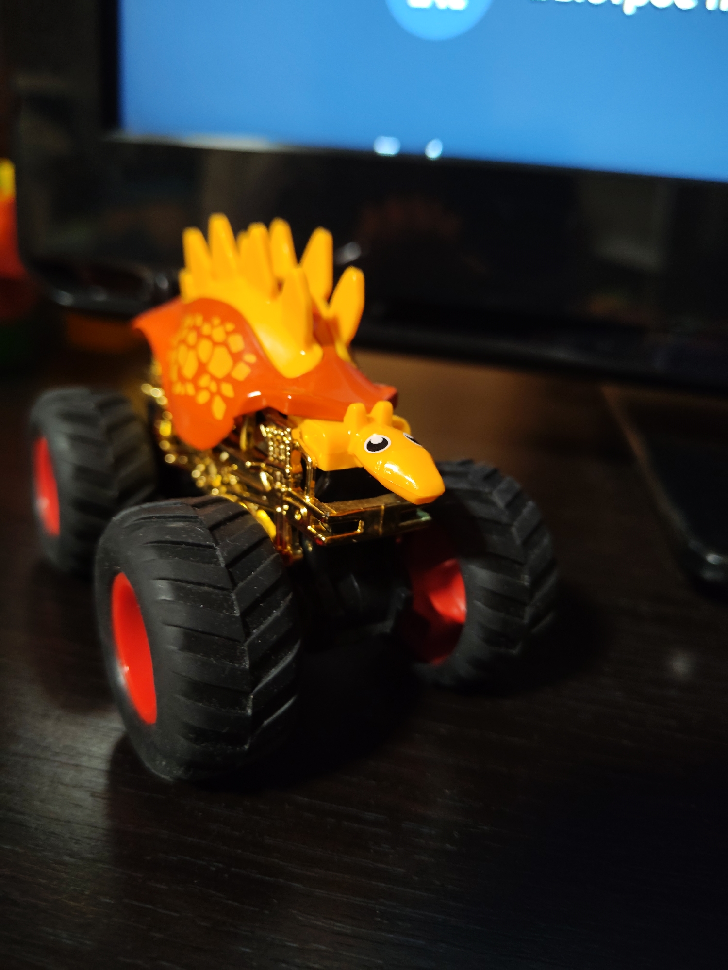 An orange and gold toy monster truck shaped like a dinosaur is shown in close-up, its large black tires with red rims resting on a dark wooden surface. The truck has a bright orange body with yellow spikes and red patterned accents, and a yellow face with black eyes. In the blurred background, a dark object, possibly a television screen displaying a blue hue, is visible. The scene is dimly lit, with a shallow depth of field emphasizing the toy truck. The overall impression is that of a playful or decorative object in a domestic setting.