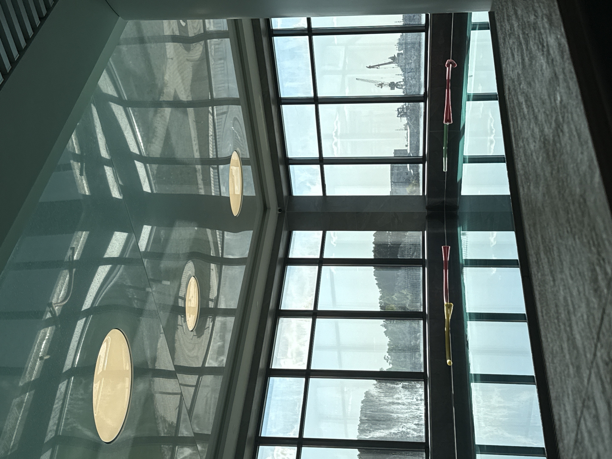 A low-angle shot indoors reveals a view through a large, multi-pane window onto an indistinct cityscape. The window consists of a grid of square panes framed in dark, possibly black, material. Visible through the window are distant structures and possibly cranes. Two decorative hanging objects, each with red and yellow components, are suspended near the right side of the window. The left side of the image consists of a reflective surface with three circular cutouts or windows. These cutouts are illuminated, casting light within them. The surrounding reflective surface shows distorted reflections of the grid-like structure of the window. A partial wall is visible on the right side of the shot with a textured, possibly gray or neutral-toned surface. The location is in the city of Unknown, Unknown.