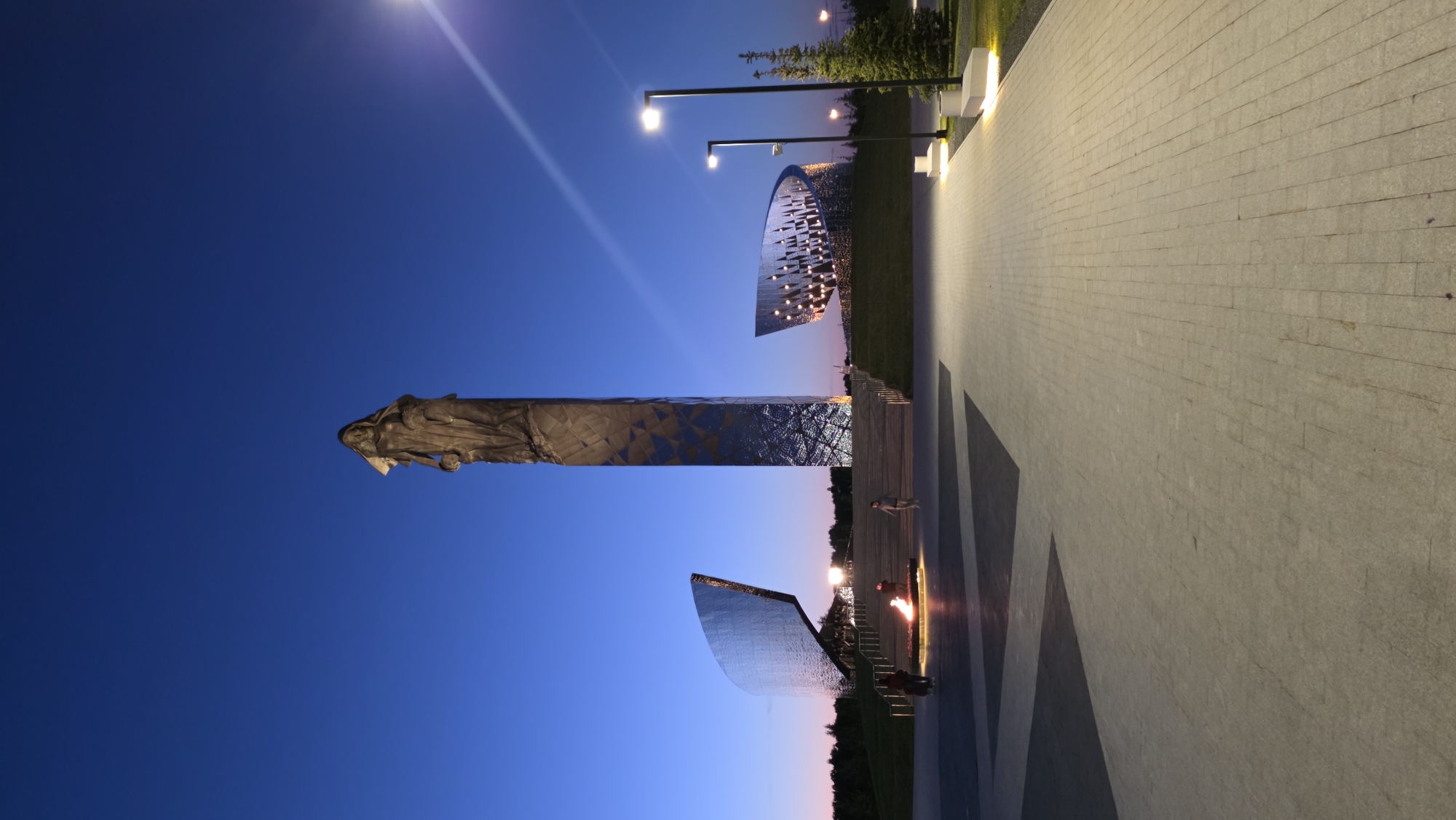 The image shows a monument complex in Unknown, Unknown, at dusk. The complex features a tall, narrow sculpture of a figure atop a similarly narrow and tall mirrored base. Additional mirrored sculptures are placed around the complex, including a semicircular arc. Several lampposts illuminate a pedestrian walkway made of light-colored stone bricks. Several people are visible in the vicinity of the complex. The sky is a gradient of blue to orange.