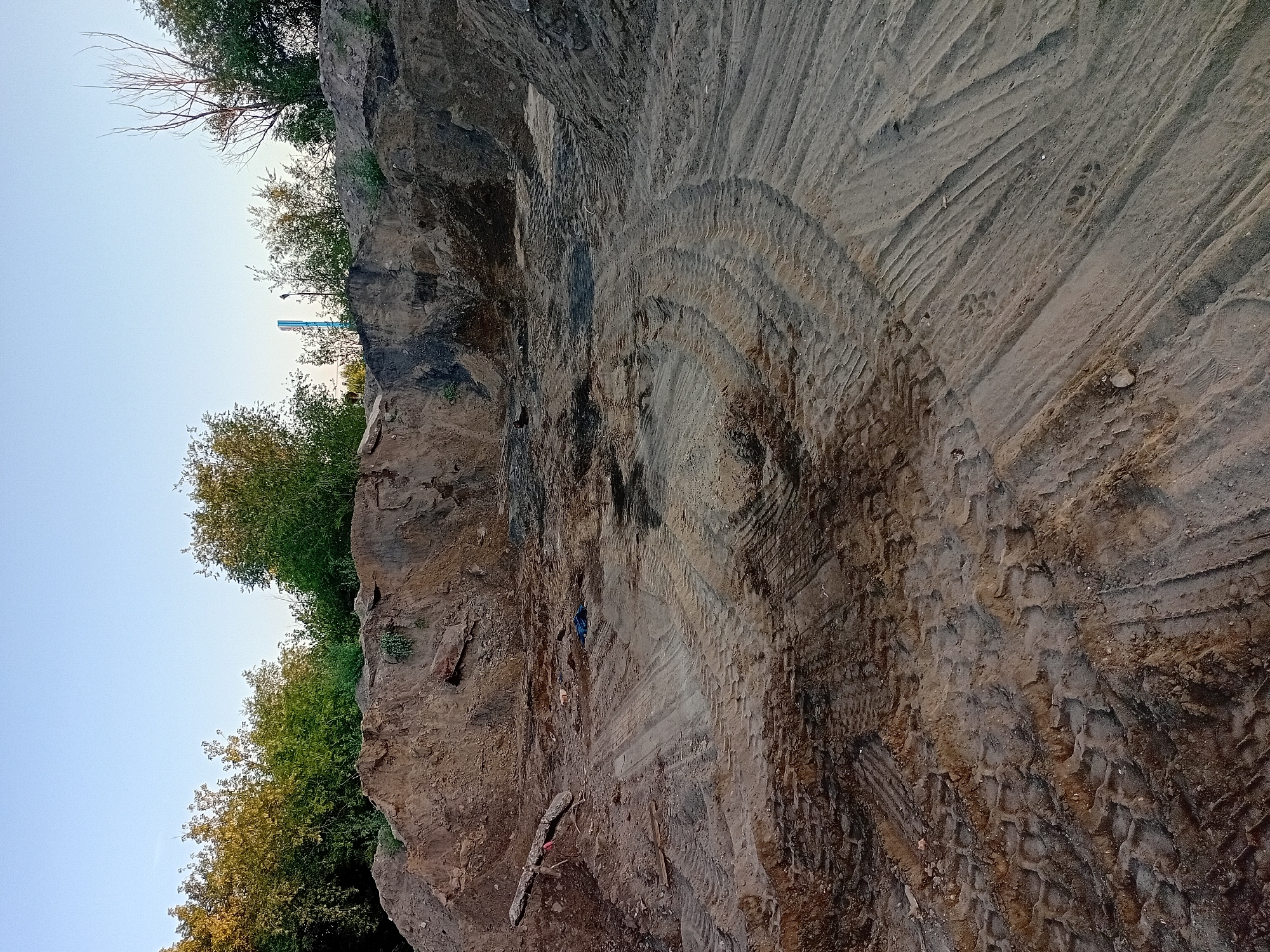 This is a photograph taken in the city of Semey, Kazakhstan. The image depicts a construction or excavation site. The foreground is dominated by a sandy, disturbed ground surface with visible tire tracks from heavy machinery, indicating recent activity. Piles of excavated soil are visible in the midground and background, creating uneven terrain. Green trees and vegetation line the upper edge of the scene, suggesting an outdoor environment. The sky above is clear and blue. There are no people or animals visible in the image. The overall impression is one of a work in progress or a site that has undergone significant earthmoving.