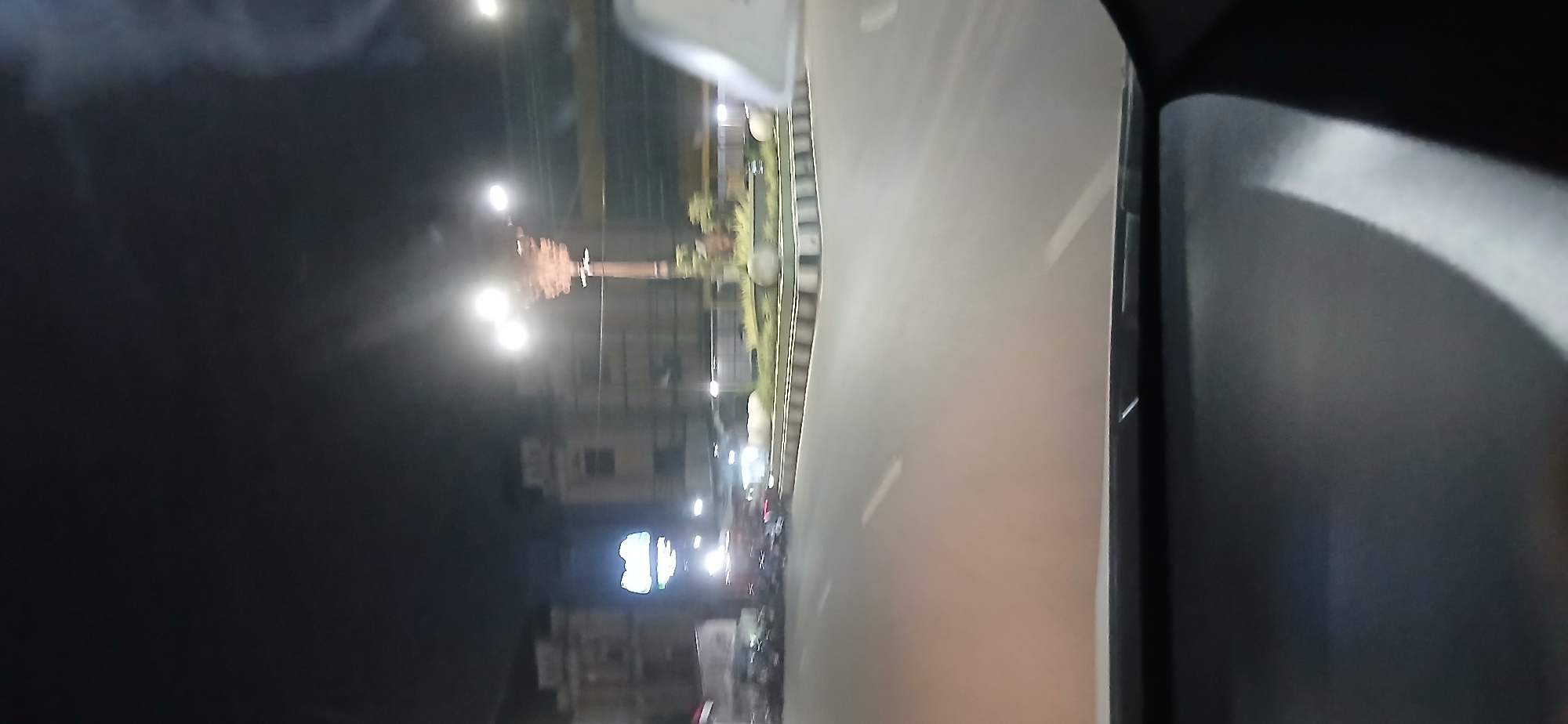 The image shows a blurred nighttime view from a vehicle, likely a car, driving through a city street in Samarinda, Indonesia. The scene is illuminated by streetlights and building lights, casting a yellowish glow on the road and surroundings. In the background, there are several multi-story buildings, some with illuminated signage. One sign appears to be a large, green, illuminated logo. A decorative street lamp with multiple bulbs is visible in the mid-ground. The road itself is paved, with white lane markings. There are some parked motorcycles on the left side of the road, and a circular island or roundabout with greenery and rocks is visible to the right, bordered by a concrete barrier. The overall impression is that of a typical urban street scene at night, likely a commercial or central area of the city.