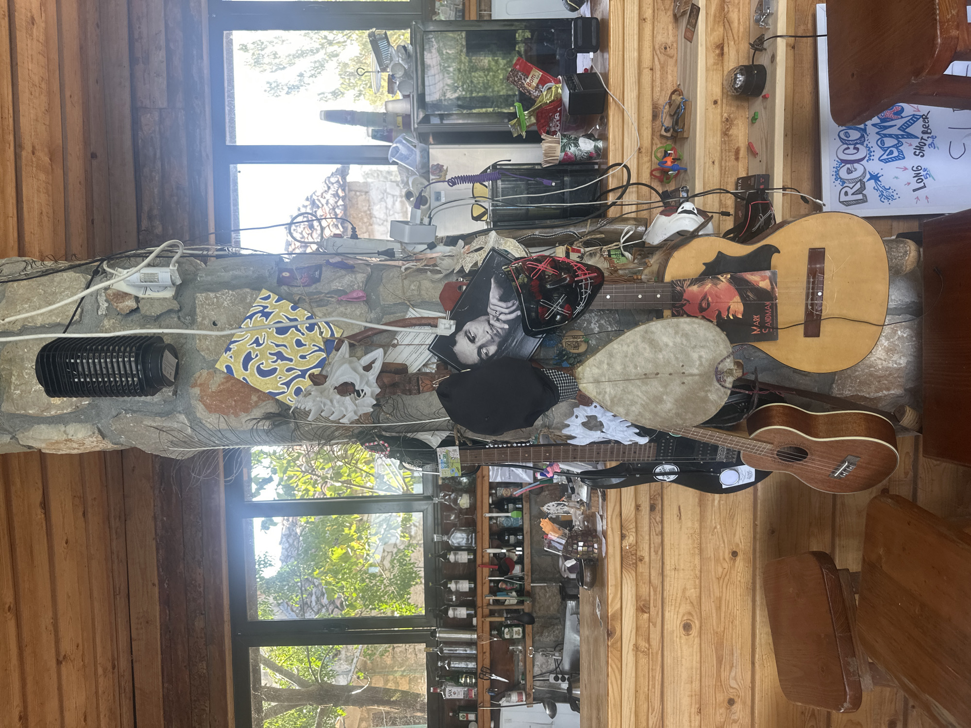 The image depicts a dimly lit, rustic interior, likely a bar or cafe, situated in Cetinje, Montenegro. A thick, stone-clad pillar dominates the center of the frame, adorned with various objects. Among these are several guitars, a ukulele, masks, and posters, including one of a man with a ring on his finger. Wires and electrical components are visible, suggesting a functional space. A wooden bar stretches across the lower part of the image, stocked with bottles of alcohol on shelves behind it. The background features large windows offering a view of greenery and what appears to be a traditional Montenegrin building. Wooden stools and tables are scattered around the foreground. The overall ambiance is eclectic and bohemian, hinting at a place with character and a love for music.