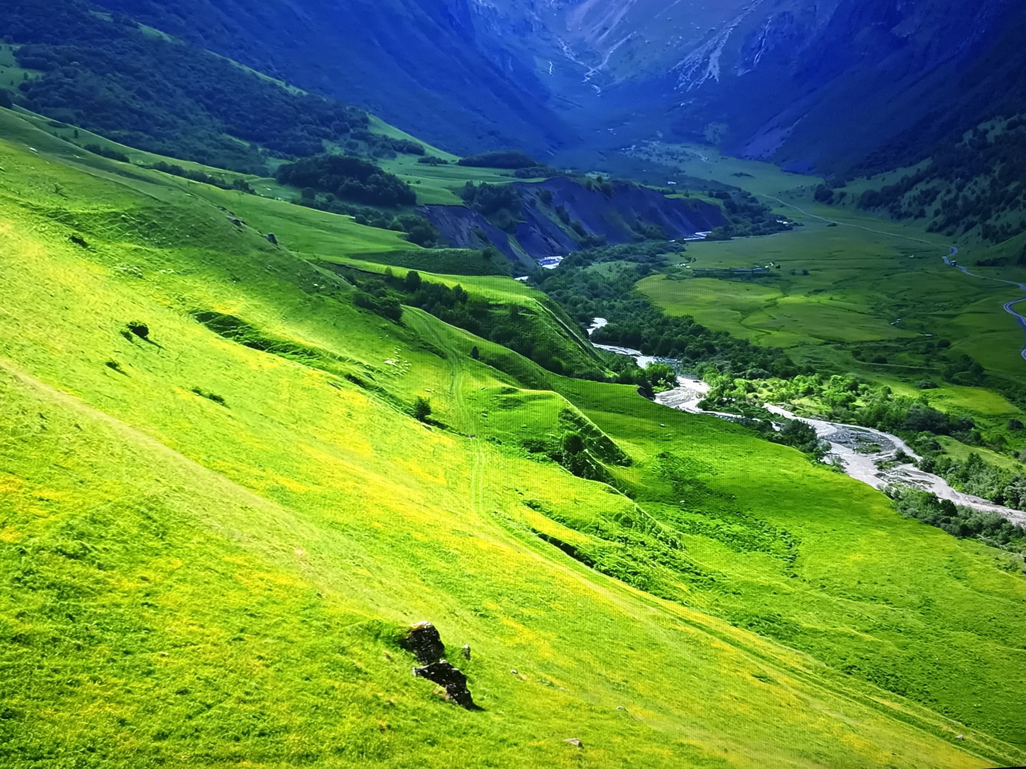 This is a landscape photograph taken from a high vantage point, overlooking a lush green valley nestled between towering mountains. The foreground is dominated by a steep, verdant hillside, bathed in bright sunlight, with patches of yellow wildflowers scattered throughout the grass. A rocky outcrop is visible on the slope. A winding river snakes through the valley floor, bordered by dense green trees. In the distance, the valley narrows and ascends towards the imposing, blue-hued mountains, their peaks softened by atmospheric haze. The scene depicts a natural, mountainous environment, likely in a rural or remote location. There are no people, animals, or man-made structures immediately apparent in the foreground or midground. The image captures a serene and expansive natural vista. The image is not related to Burgas, Bulgaria.