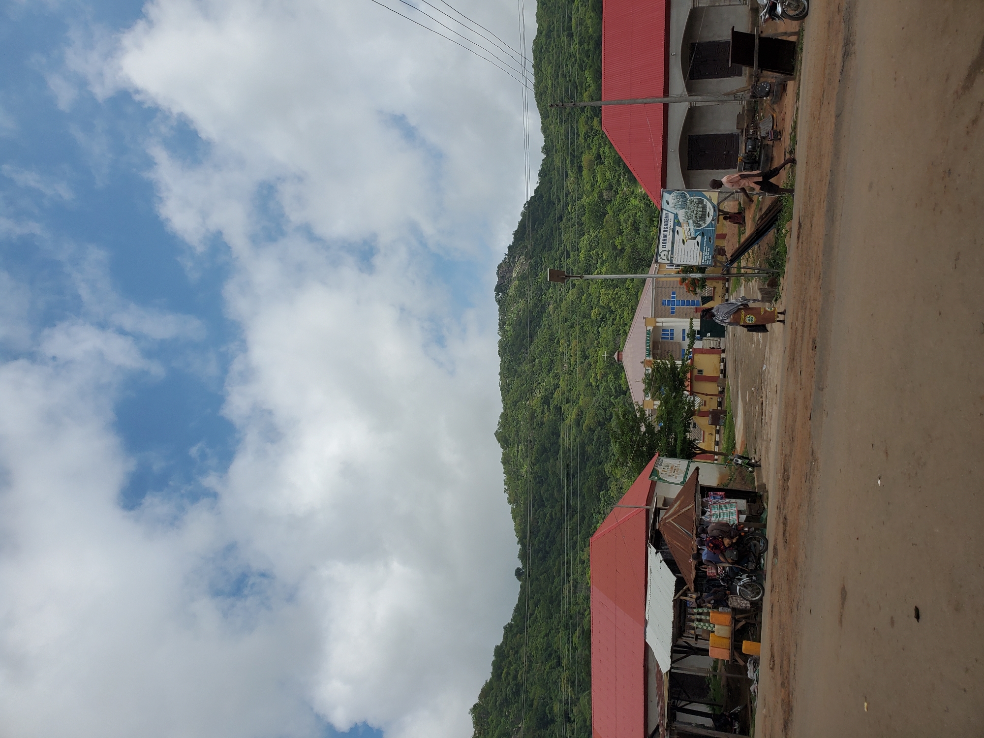 This image captures a vibrant street scene in Mararaba, Nigeria, under an overcast but bright daytime sky. The top portion of the frame shows a blue sky dotted with white clouds.
To the left, a large, lush green mountain, densely covered in trees, dominates the background, giving the scene a natural, rugged backdrop. Along the right side of the image, an unpaved, dusty road stretches into the distance, with several people engaged in daily activities.
On the right side of the road, various structures line the street. A prominent building with a red corrugated iron roof and dark, arched entrances is visible. Further along, another building with a lighter blue and white facade is seen, featuring a large blue and white billboard that clearly displays "ELOHIM ACADEMY," indicating an educational institution. A street light pole stands nearby. In the foreground on the right, a bustling market stall with a rusty corrugated iron roof offers goods, including several yellow jerrycans, and attracts a small group of people. Two motorcycles are parked near the market area and further down the street.
Several individuals are present: one person in a light-colored shirt walks past the "Elohim Academy" billboard, and another individual in a patterned garment stands further down the road. A group of people are gathered around the market stall, suggesting active commercial exchange. Multiple power lines traverse the sky above the street and buildings. The scene depicts typical daily life in a semi-urban setting, characterized by local commerce and movement of people.