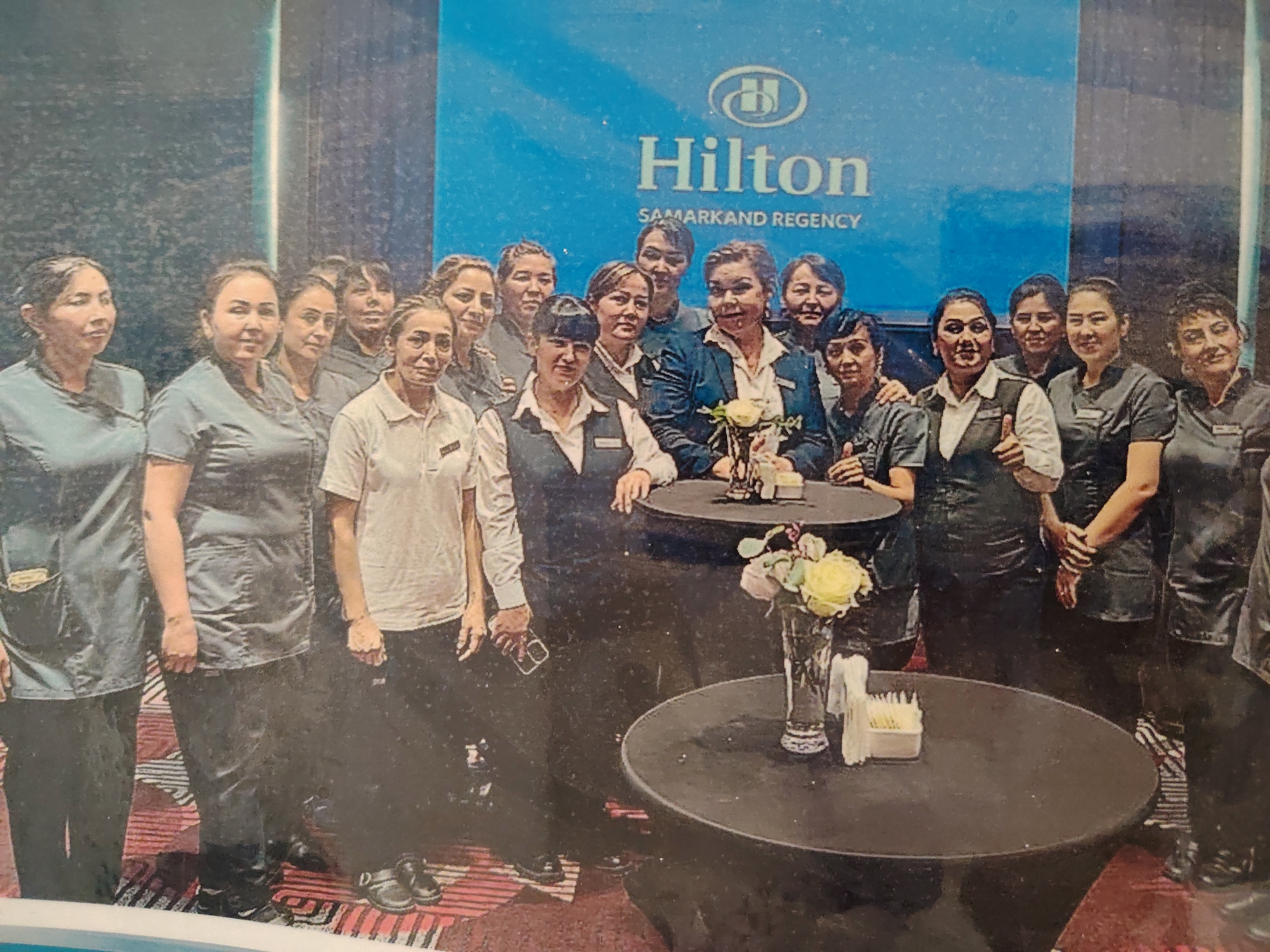 In Konigil, Uzbekistan, a group of fifteen individuals, predominantly women, pose for a photograph in front of a large blue backdrop with the "Hilton Samarkand Regency" logo. The group appears to be employees, as many are wearing similar dark uniforms. The individuals are arranged in two rows, with the front row standing and the back row slightly behind. Some individuals are smiling, while others have neutral expressions. The setting suggests an indoor event or gathering, possibly at the Hilton hotel. There are two round cocktail tables in the foreground, one of which has a vase with flowers and a small container with napkins. The lighting is moderate, and the overall image quality suggests it may be a scanned photograph.