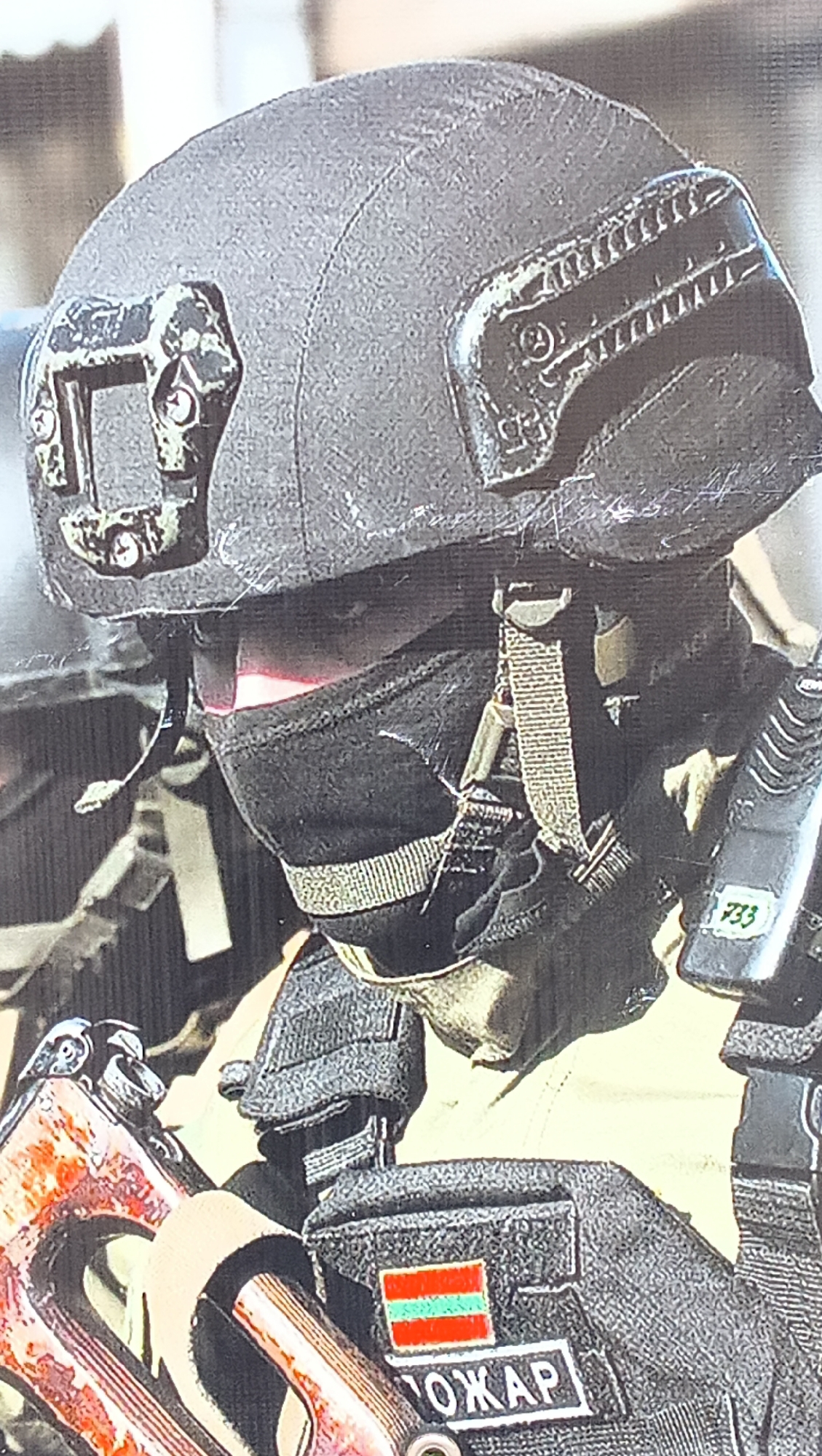 A close-up, chest-up portrait shows an individual in tactical gear, likely outdoors in daylight. The subject wears a dark grey or black ballistic helmet with side-mounted accessory rails and a black balaclava or face covering that conceals most of the face, with a subtle red glow visible where the eyes would be, possibly from eyewear or a reflection.
The individual is equipped with a dark tactical vest or plate carrier. Attached to the right side of the vest is a radio or communication device marked with the numbers "733". A distinctive flag patch on the vest features horizontal stripes of red, green, and red, which is the flag of Transnistria. Below this flag, a name patch displays the Cyrillic text "ПОЖАР", which translates to "POZHAR" or "Fire".
In the lower-left foreground, a portion of a rifle is visible, identifiable by its reddish-brown foregrip and receiver, suggesting an AK-pattern weapon. The background is blurred, emphasizing the subject and their equipment, indicative of a military or security professional in Tiraspol, Moldova (Transnistria). The scene conveys a sense of readiness and operational posture.