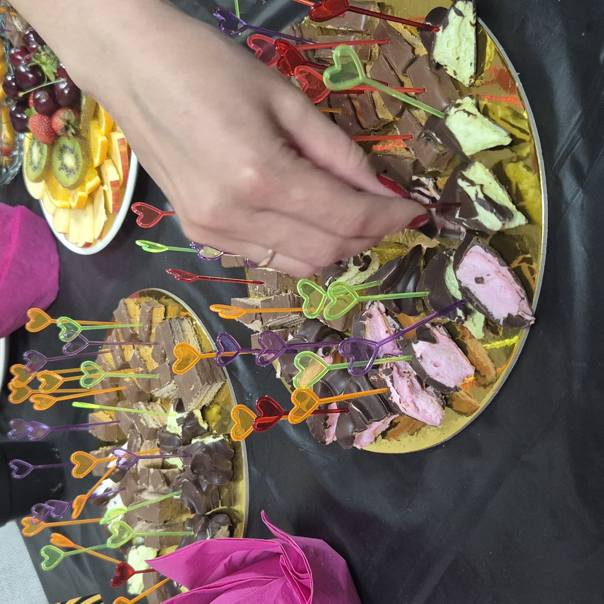 The image shows a close-up of a dessert platter with chocolate-covered sweets, likely at a party. The platter is gold and round, filled with various bite-sized treats. Many of the sweets are skewered with colorful plastic heart-shaped picks in shades of orange, purple, green, and red. Some of the sweets appear to be layered wafers with a light-colored filling, while others are covered in chocolate with a pink or green marshmallow-like filling. A person's hand, with red nail polish, is reaching in to pick up one of the sweets. To the left, a folded pink napkin is visible. In the background, a plate of sliced fruit including oranges, kiwi, strawberries, and cherries can be seen, along with a pink fabric covering. The scene is set on a black tablecloth. There is no information in the image to indicate the location is Kokshetau, Kazakhstan.