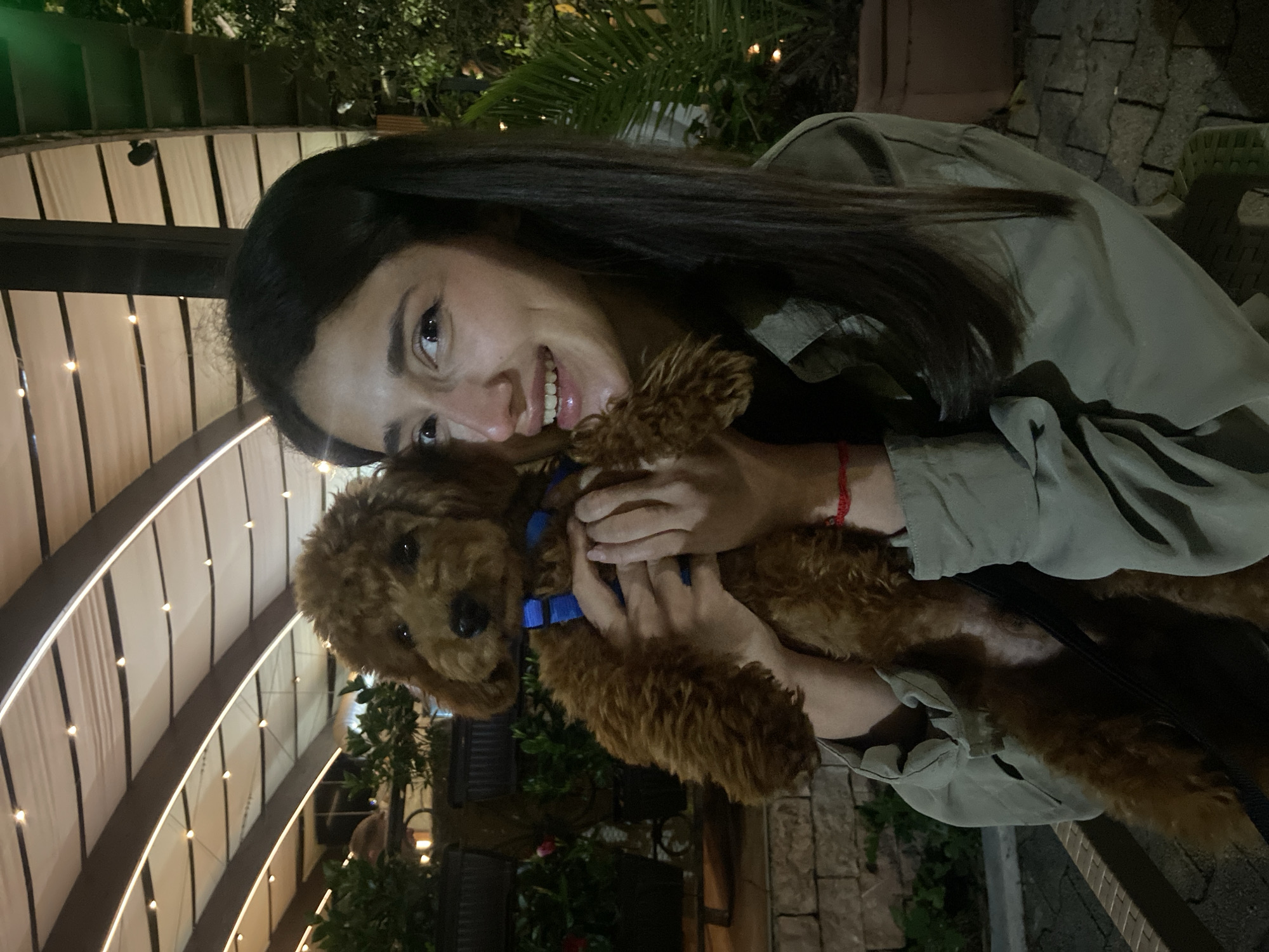 A young woman with long, dark hair is holding a small, brown poodle dog in her arms. The dog is wearing a blue collar. The woman is smiling and looking at the camera. They are outdoors, seemingly at night, with ambient lighting from string lights visible in the background and some foliage. The location is likely in Bar, Montenegro, based on inference of the origin request. The woman appears to be wearing a light-colored jacket. The background consists of a covered area with string lights and greenery.