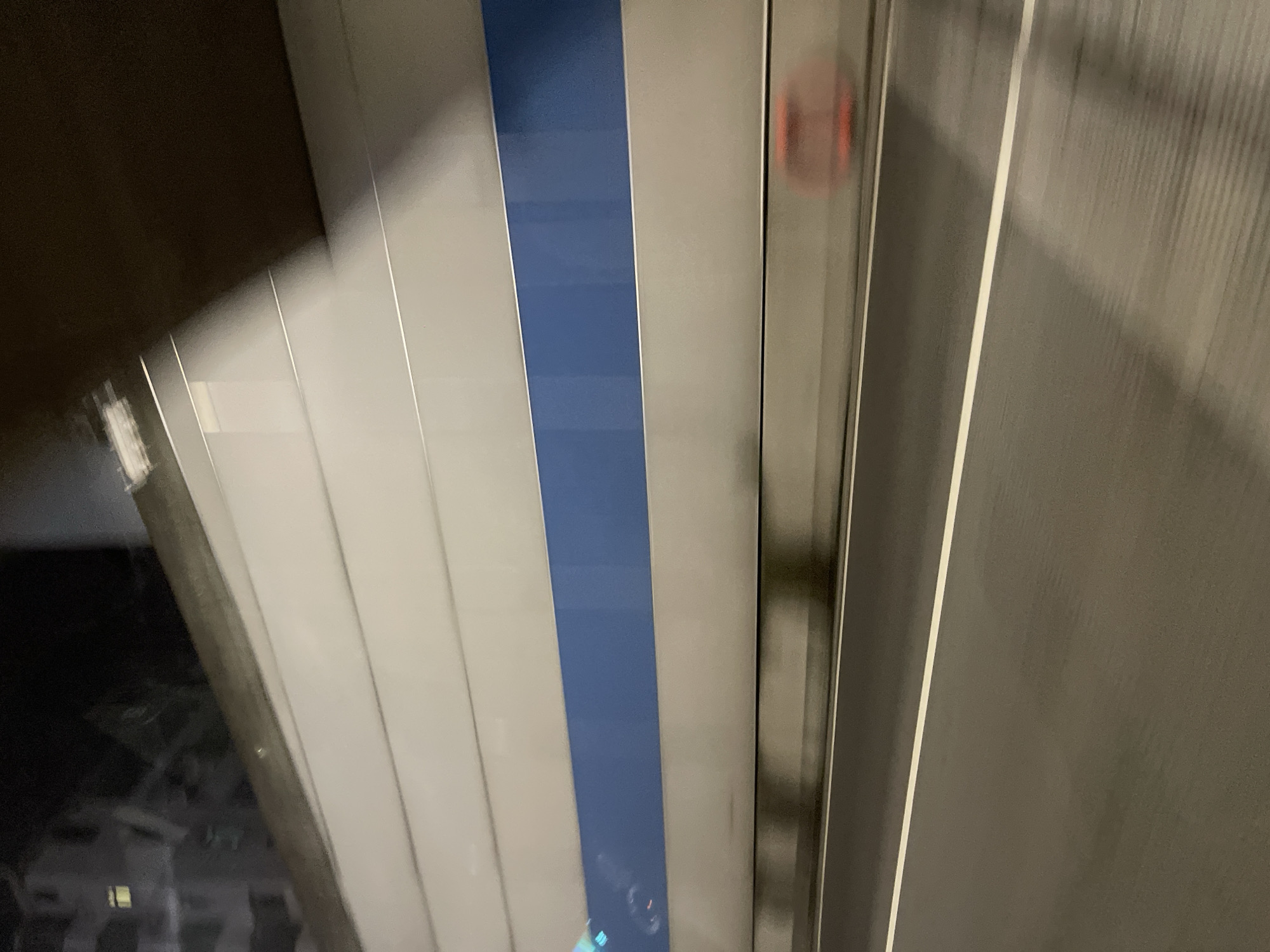 The image depicts a view from inside a vehicle traveling through a tunnel or underpass, likely in Panyu District, China, given the context. The scene is characterized by motion blur, indicating movement. The primary elements visible are the road surface, the tunnel walls, and the ceiling. The road is asphalt, marked with a white line indicating a lane. The tunnel walls are constructed from horizontal panels, predominantly light gray, with a horizontal band of dark blue in the upper section. Above the walls, a portion of the tunnel ceiling is visible, appearing dark and curved. In the distance, on the right side of the frame, a blurred, reddish-orange object is visible near the tunnel wall, possibly a warning light or a piece of equipment. The left side of the image shows a reflection of buildings and lights, suggesting the tunnel is near an urban area. The overall impression is one of a functional, utilitarian infrastructure environment within a city.