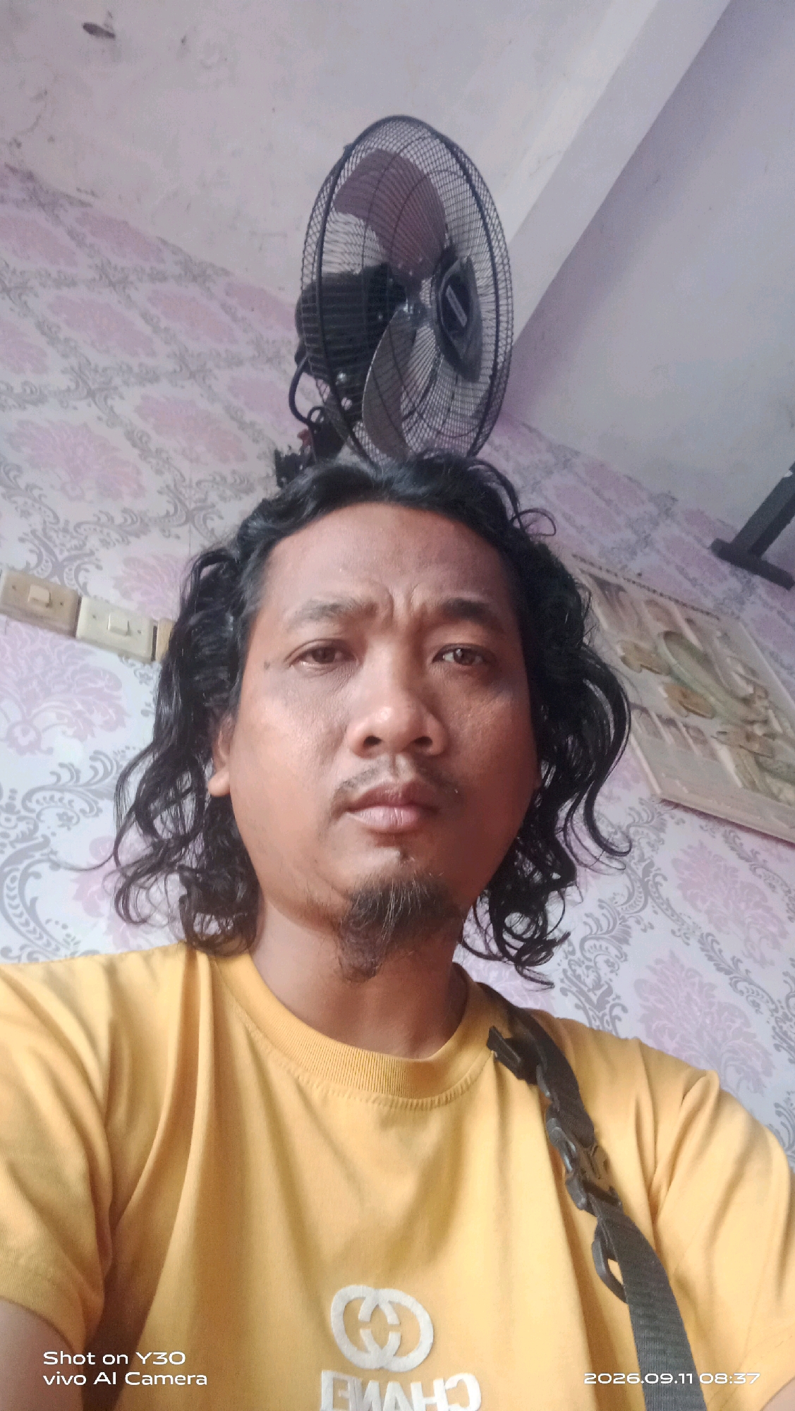 A medium shot selfie shows a man with long, dark, curly hair and a goatee, looking directly at the camera. He is wearing a yellow t-shirt with a white logo on the chest, which appears to be a stylized "CC" or interlocking rings, and partially visible text below it. A black strap is visible across his right shoulder. The background consists of a patterned wallpaper in shades of pink and gray, and an electrical fan is mounted on the wall behind and above him. To the right, a framed picture or artwork is hanging on the wall. The image also contains text overlays in the bottom left corner: "Shot on Y30 vivo AI Camera" and in the bottom right corner: "2026.09.11 08:37". This image was taken in Munggugianti, Indonesia.