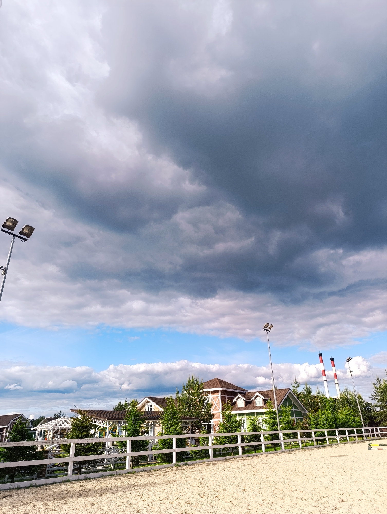 The image shows an outdoor equestrian arena in Ivanovskoe, Russia. The arena is enclosed by a white fence and contains a sand surface. Beyond the fence, several buildings are visible, including structures with brown roofs and light-colored walls. The buildings are partially obscured by trees. In the background, two tall industrial chimneys with red and white stripes can be seen. Lighting poles are positioned around the arena. The sky is overcast with a mix of dark gray storm clouds and patches of blue sky.