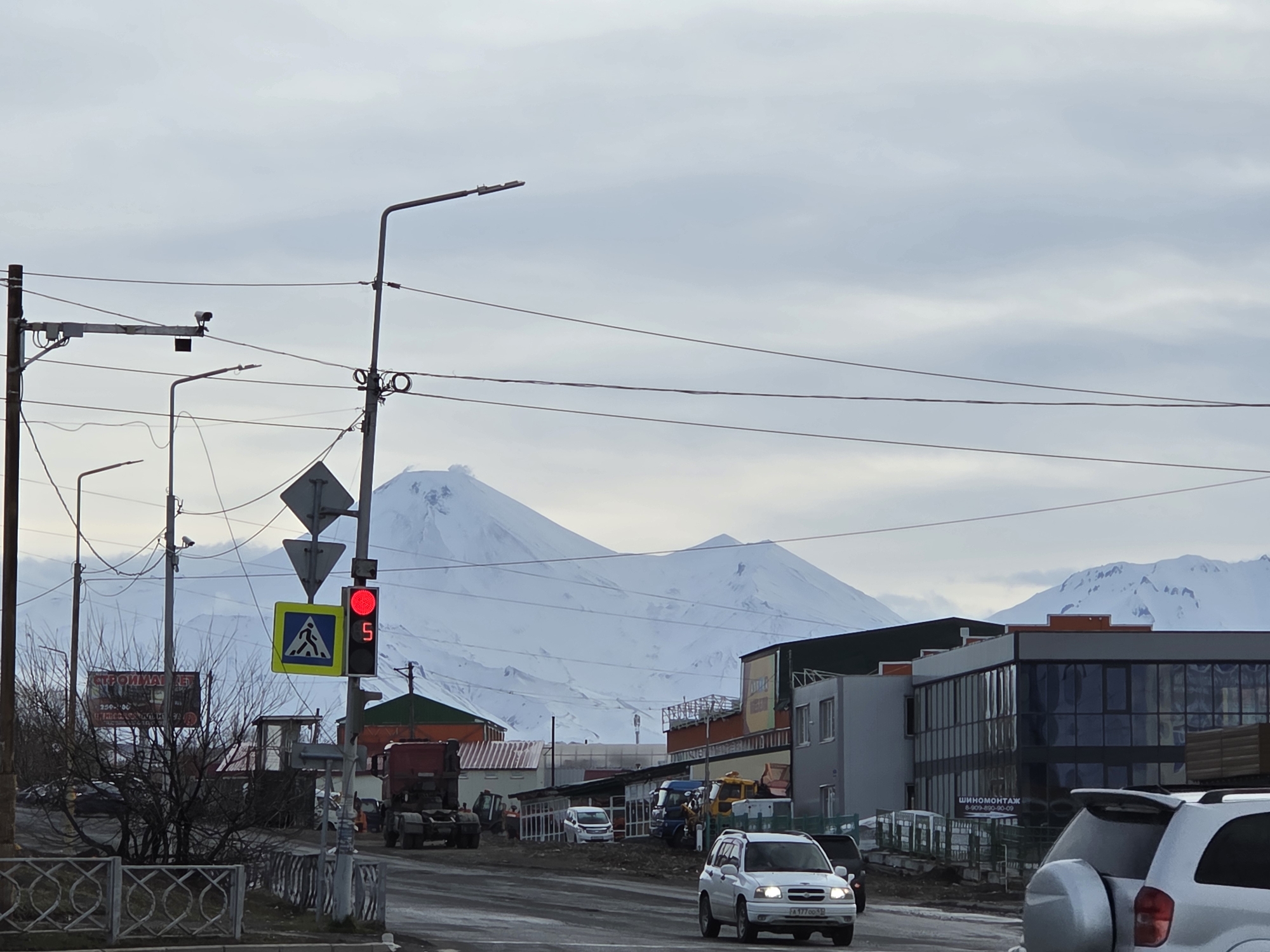 The image shows a street scene in an unknown city. The weather appears overcast. In the background, there are snow-covered mountains. In the foreground, there's a road with cars driving on it. There's a pedestrian crossing with a traffic light displaying a red light. Buildings of various sizes and designs line the side of the road. Power lines and streetlights are visible overhead. A billboard with Russian text is present. There are also road signs and construction equipment in the vicinity. The scene appears to be a mix of residential and commercial areas.