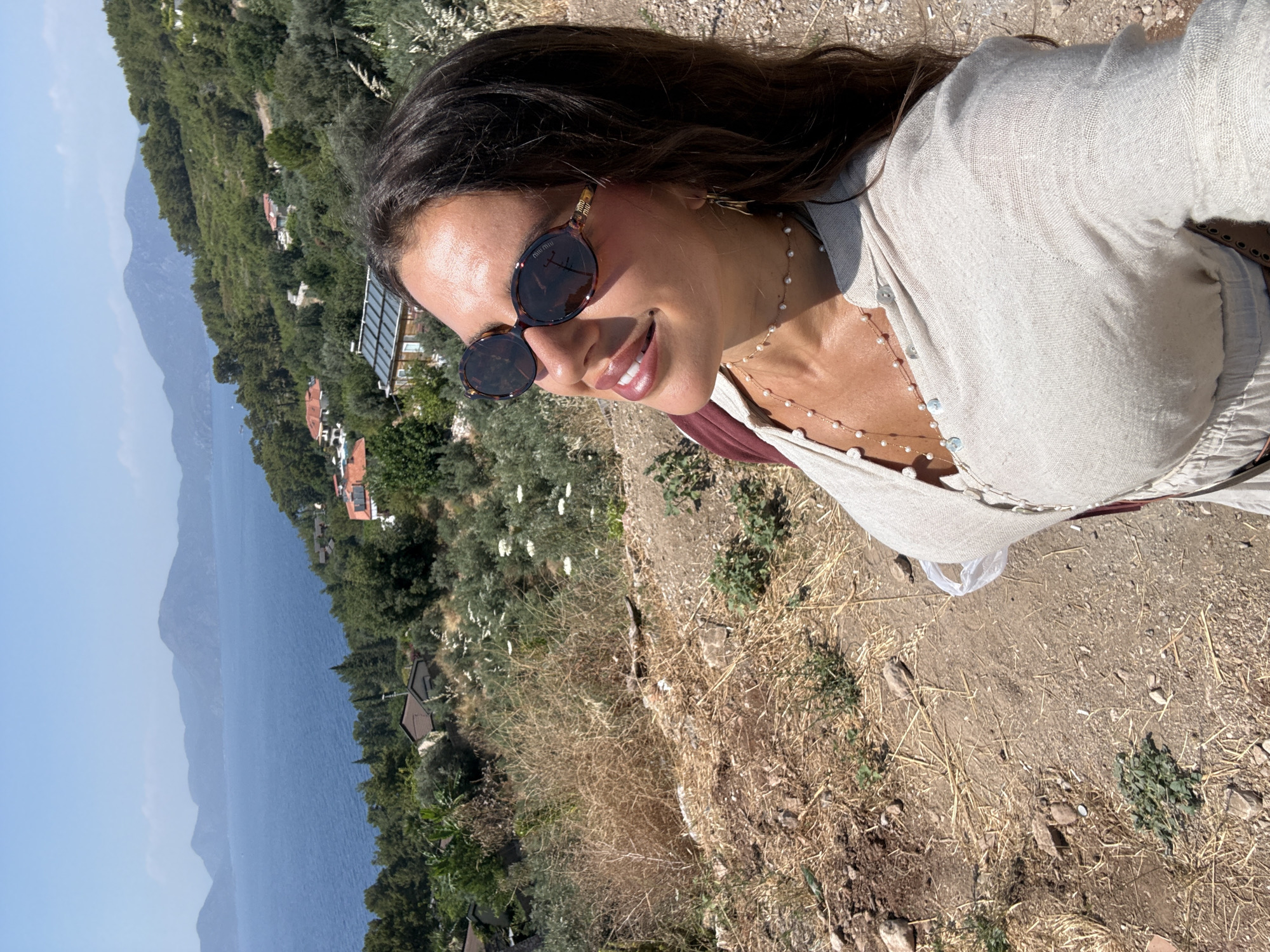 A woman with long, dark hair is taking a selfie outdoors. She is wearing sunglasses with tortoise-shell frames and a light-colored, woven shirt. A delicate pearl necklace is visible around her neck. She is smiling and looking to her right. The background reveals a scenic view of a body of water, possibly the sea, with distant mountains under a clear blue sky. The terrain in the foreground is dry and rocky, with some sparse vegetation. Scattered amongst the greenery on the hillside are several small buildings with reddish-brown roofs, suggesting a village or resort. The lighting indicates it is daytime, likely late morning or afternoon, with bright sunlight. The overall mood appears relaxed and contented. The location is Fethiye, Turkey.