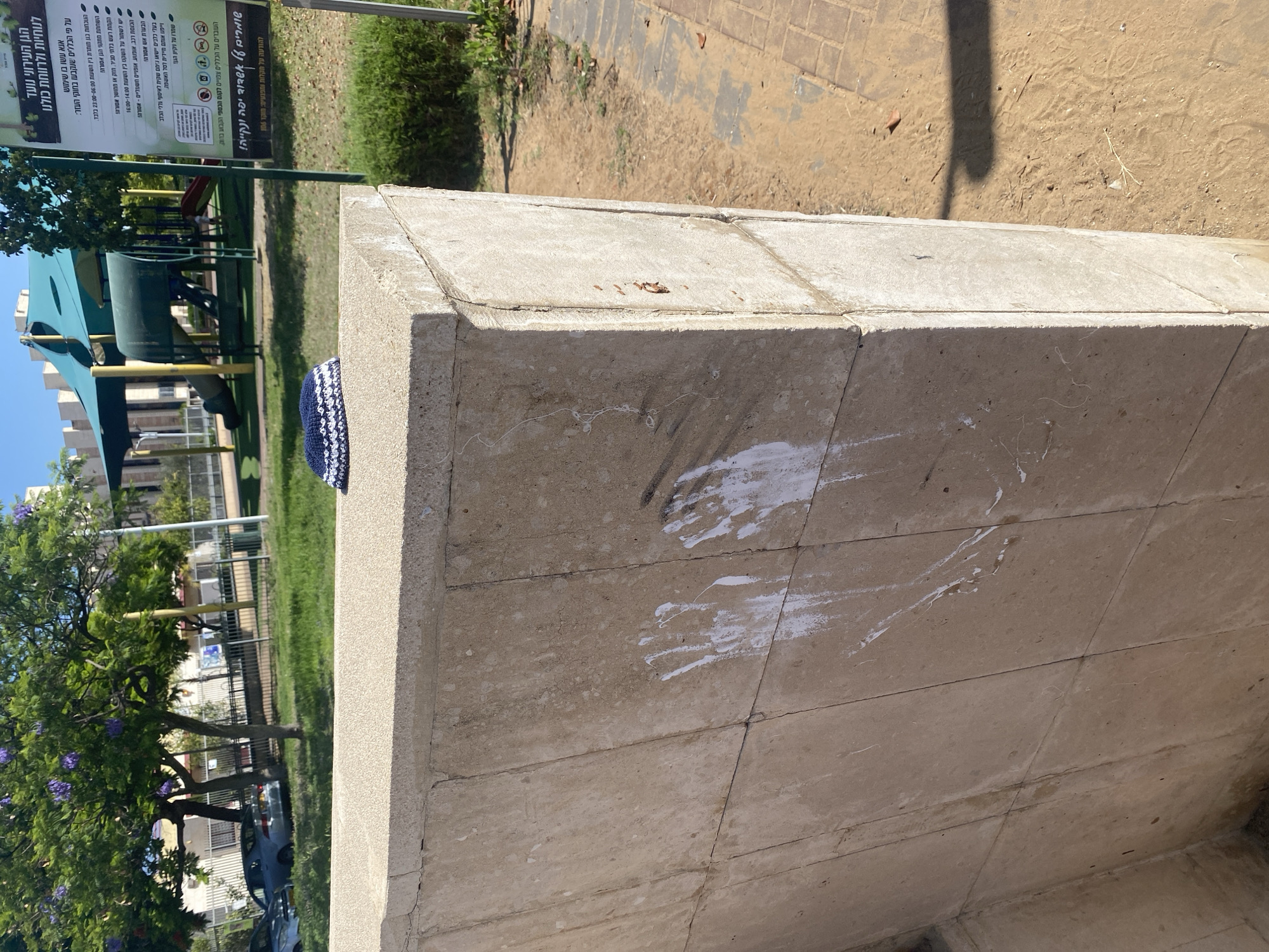 An outdoor scene in Ashdod, Israel, shows a light-colored, textured concrete structure in the foreground. On top of this structure rests a small, white and blue knitted cap. The concrete surface of the structure has several white handprints and smudges, appearing to be paint. In the background, a park area is visible with a playground featuring slides and climbing structures under a green canopy. Trees with purple flowers are also present, along with cars parked on a street and a fence. A sign with Hebrew text stands near the playground. The ground is a mix of dirt and paved brick pathways. The overall lighting suggests a sunny day.