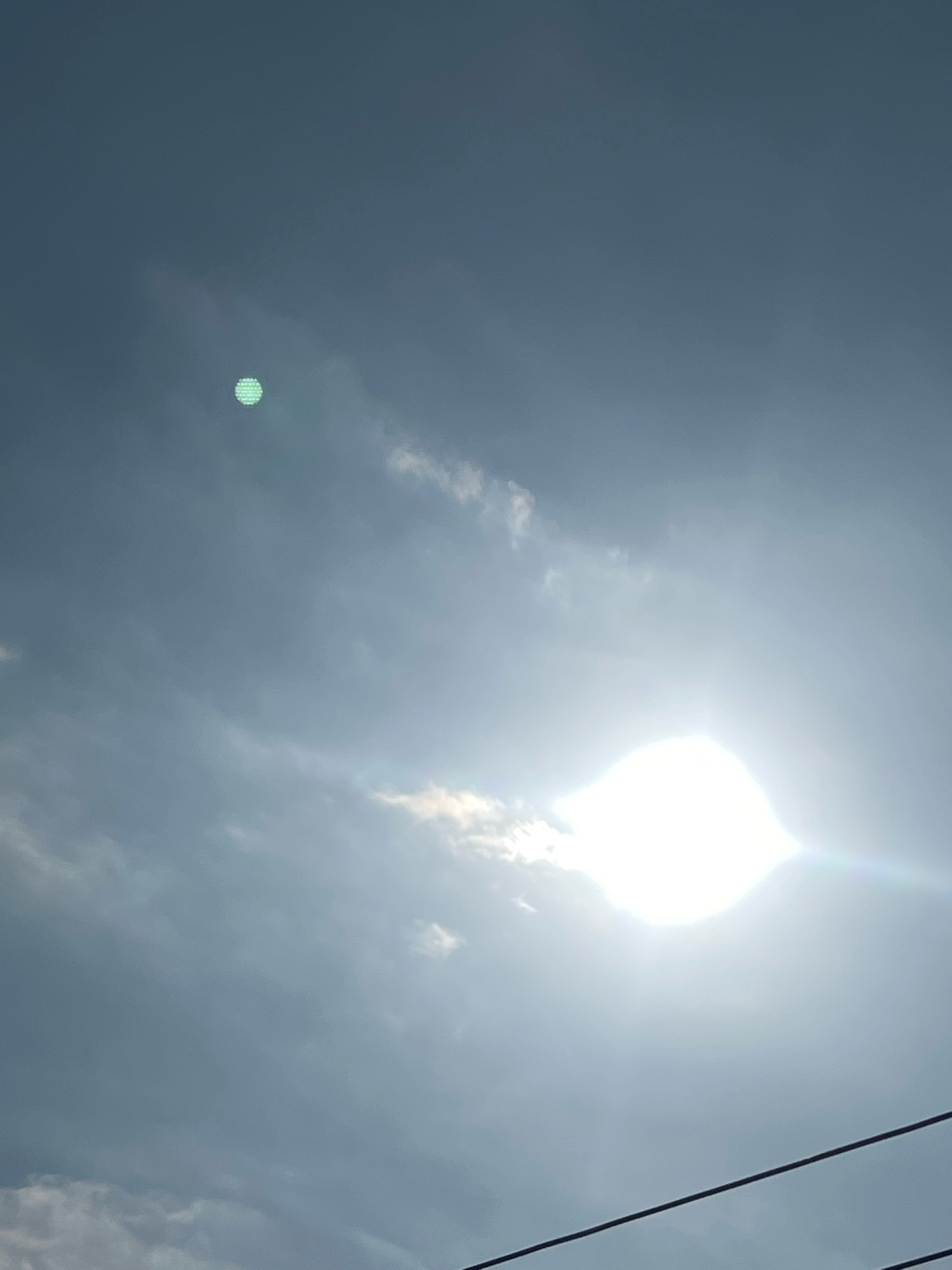 The image shows a hazy sky with clouds and the sun prominently featured on the right side, creating lens flare. In the upper left quadrant of the sky, there is a distinct circular object with a green, patterned surface. The foreground contains the top of power lines. The location is the city of Denpasar, Indonesia.