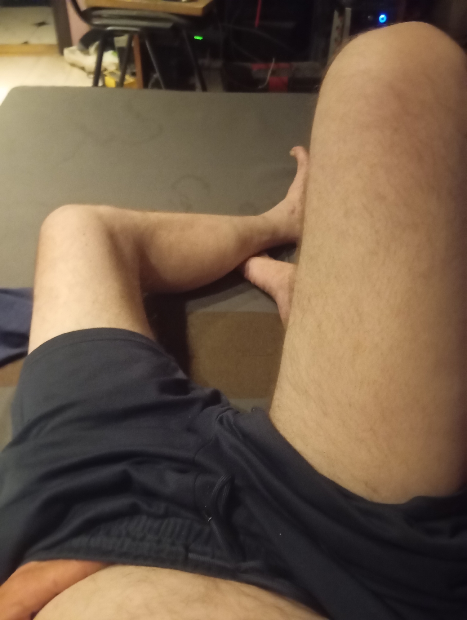 The image shows a close-up view of a person's legs and torso. The person is wearing dark blue athletic shorts. Their legs are slightly hairy, and they are resting on a dark grey surface. The background is blurred, but it appears to be an indoor setting with some furniture and electronics visible. The lighting is dim, suggesting it might be evening or night. There is no visible text in the image. The overall impression is of a casual, relaxed moment.