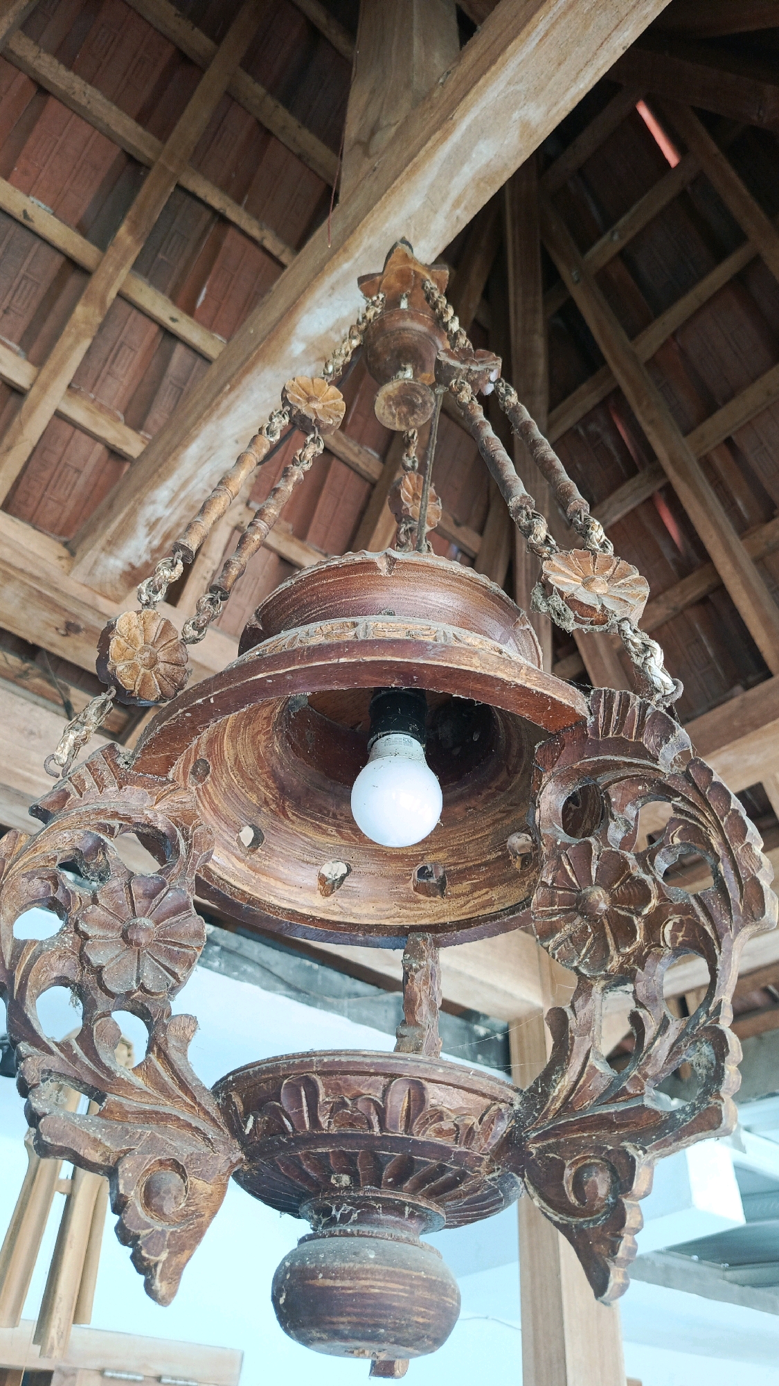 The image shows an ornate, wooden chandelier hanging from a thatched roof structure. The chandelier is carved with floral motifs and is suspended by chains. A single lightbulb is visible within the central part of the chandelier. The background reveals the interior of a building with exposed wooden beams and terracotta tiles forming the roof. The overall scene suggests a traditional or rustic setting in Babadan, Indonesia.